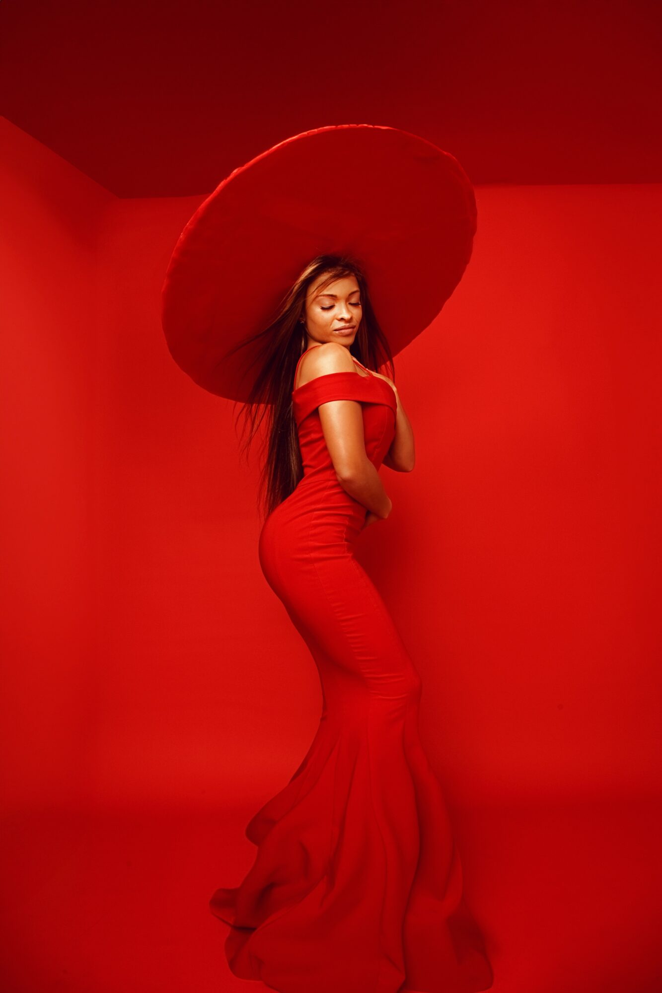 Woman in red dress holding large red umbrella against red background, looking over shoulder, long hair, elegant pose.