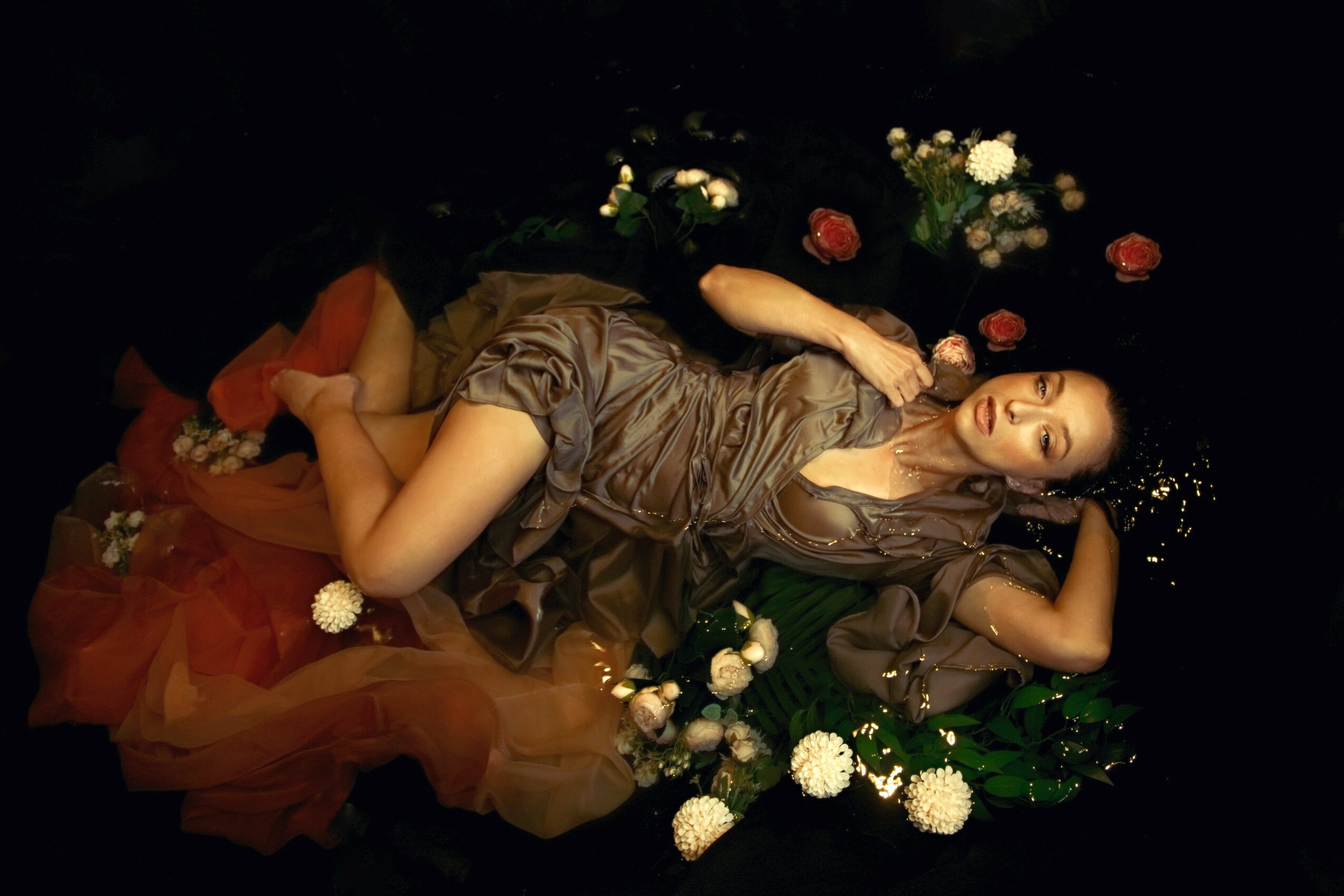Woman lying on her side surrounded by flowers, wearing a brown dress, with her eyes closed and arms resting on her head.