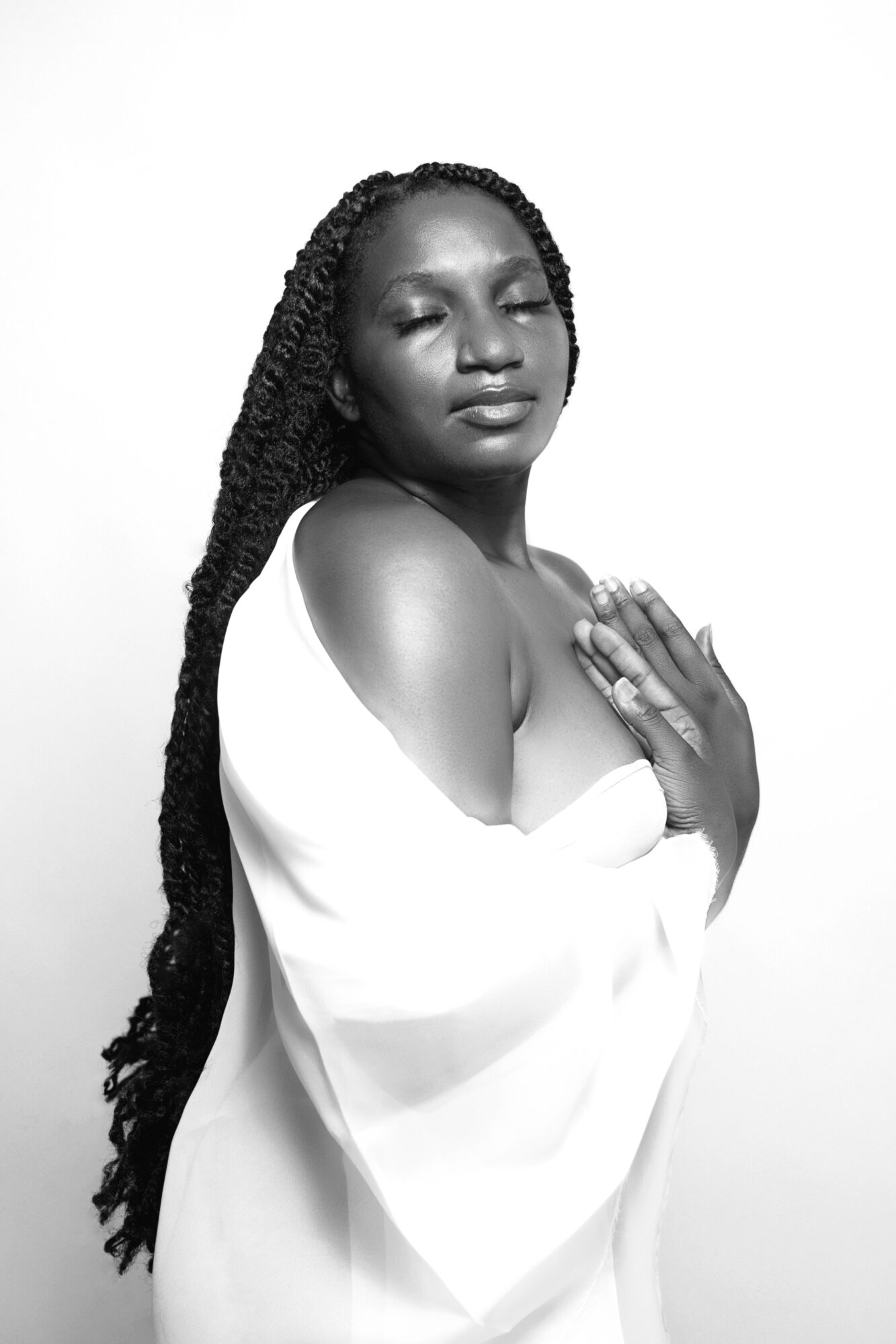Woman with long braided hair, eyes closed, hands on chest, wearing a white off-shoulder garment, against a plain background.