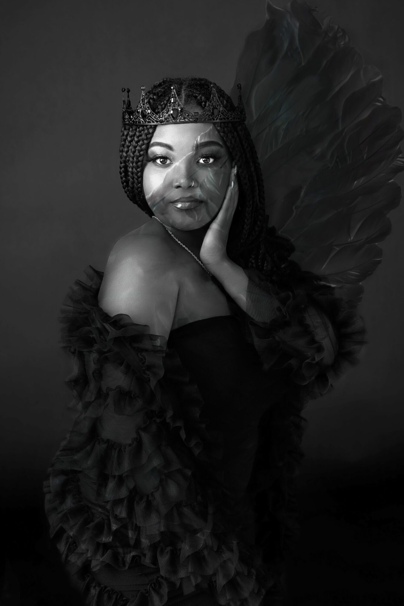 Woman with braided hair, wearing a crown and feathered outfit, posing with hand on face, in black and white.