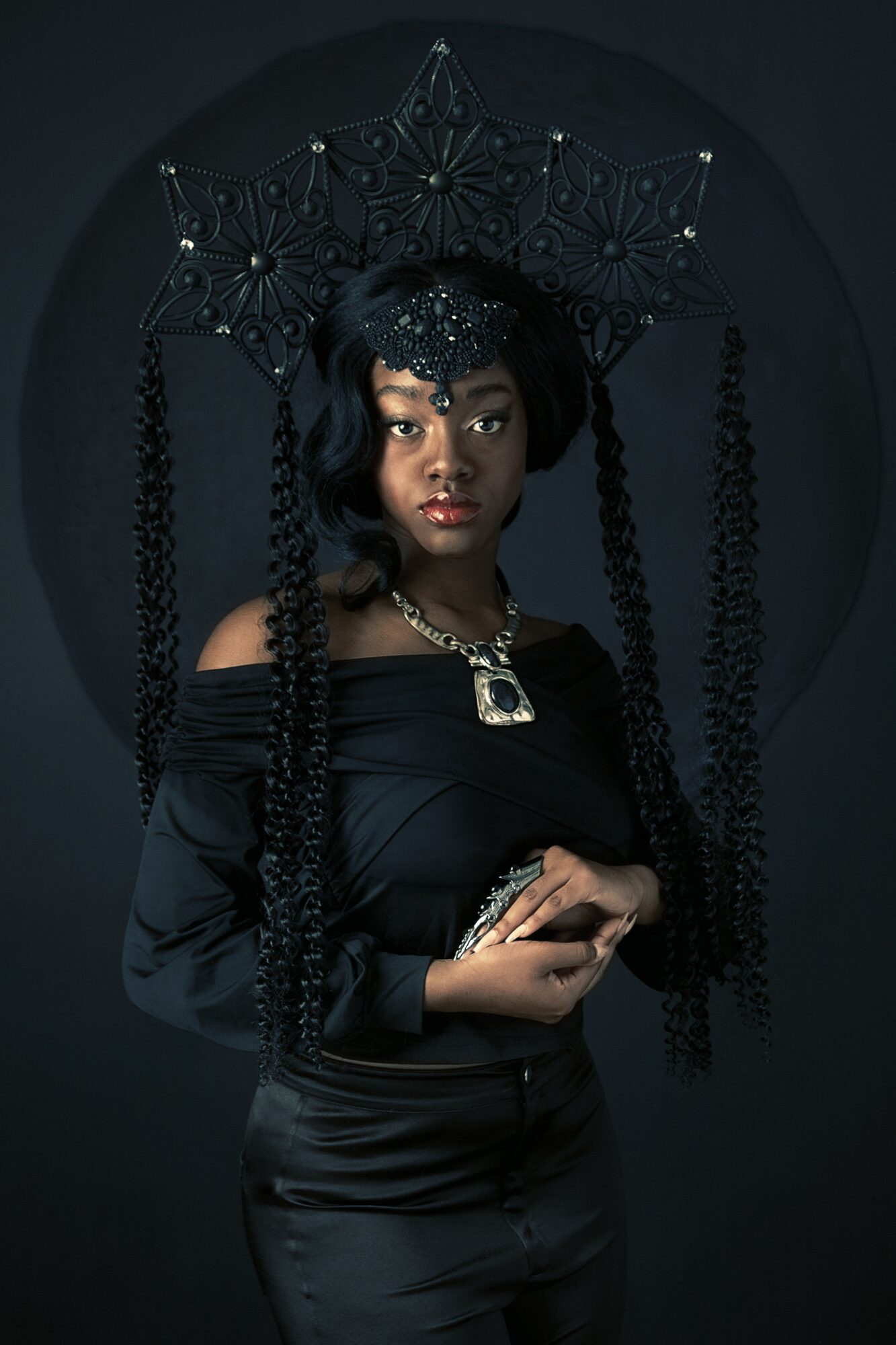 Woman with elaborate black headpiece and jewelry, wearing a black off-shoulder top, against dark background.