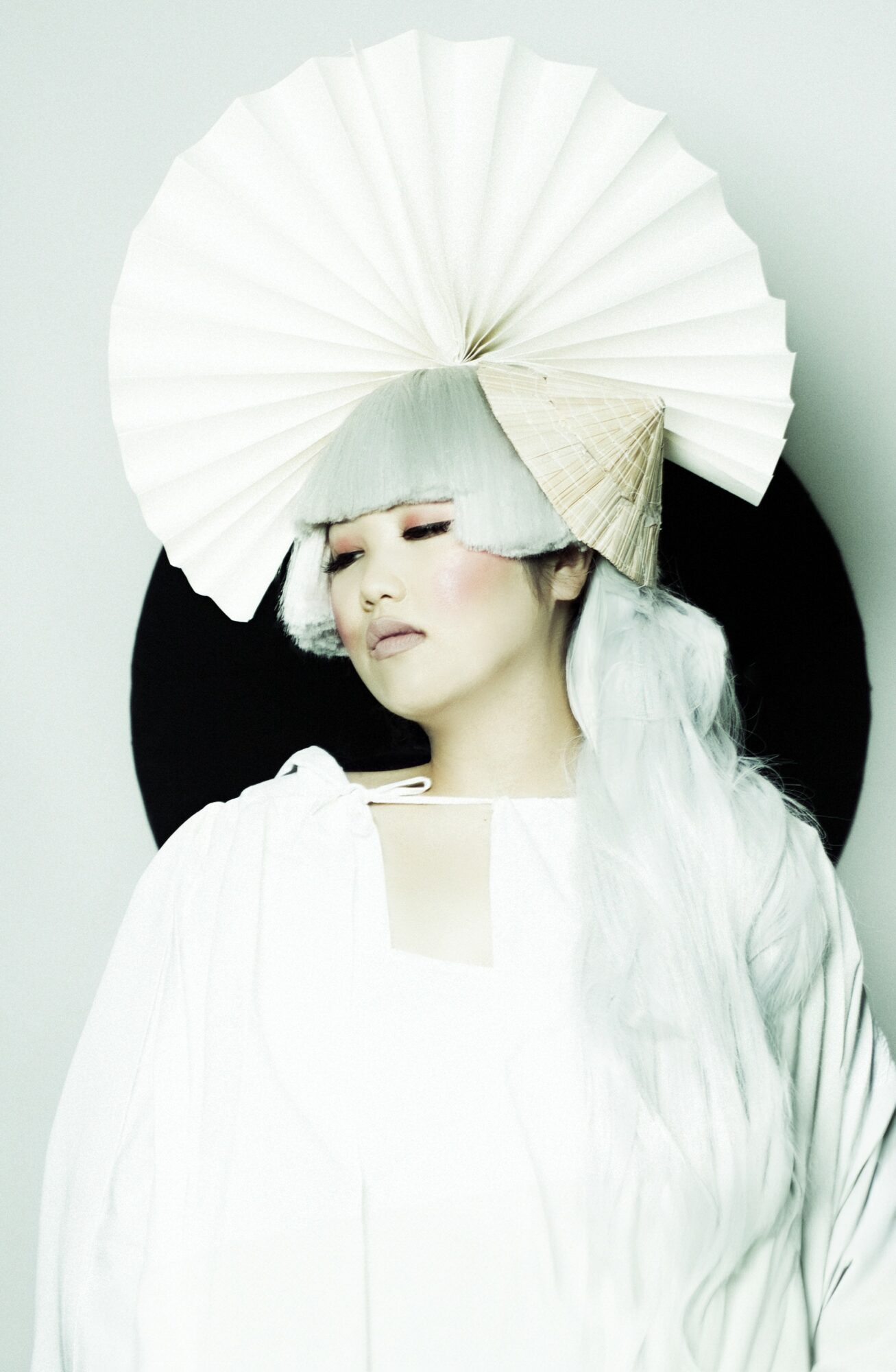 Person with elaborate white headpiece and flowing white clothing, eyes closed, against a black background.