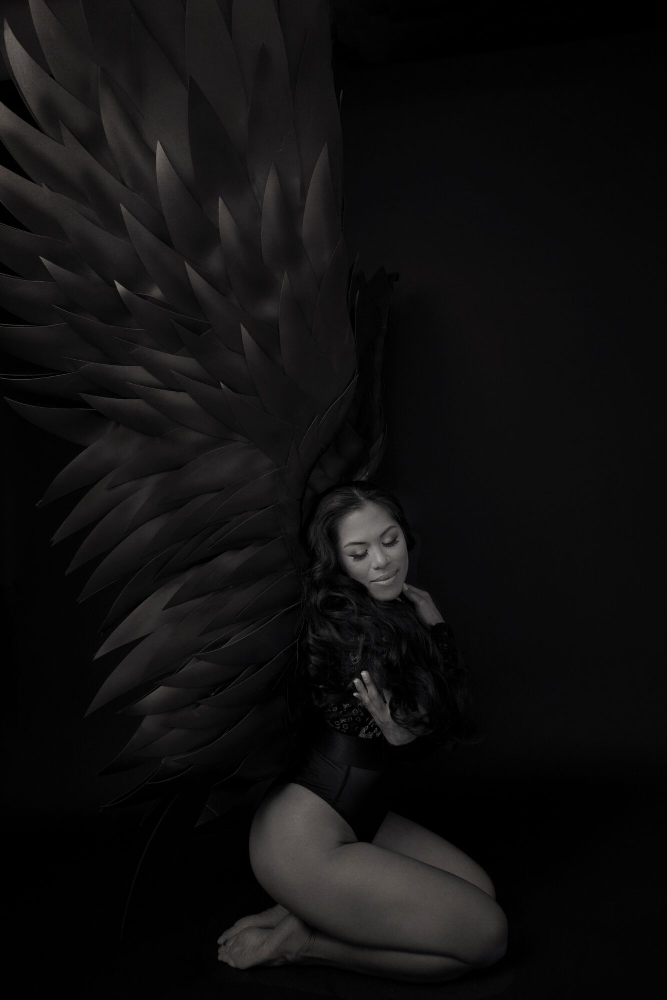 Woman with large dark wings, sitting on the floor with eyes closed, in a dark setting.