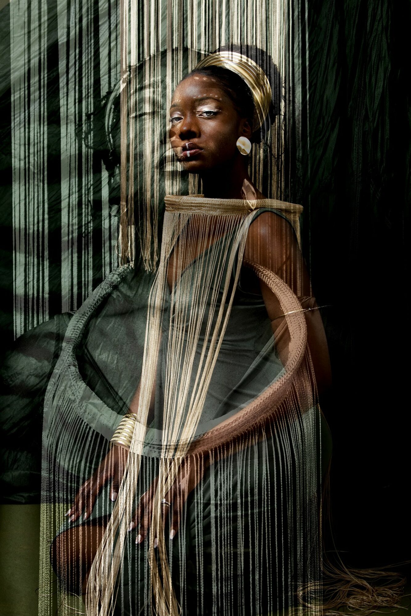 Portrait of a woman with a headwrap, sitting with her hands on her lap, against a dark background with vertical lines.