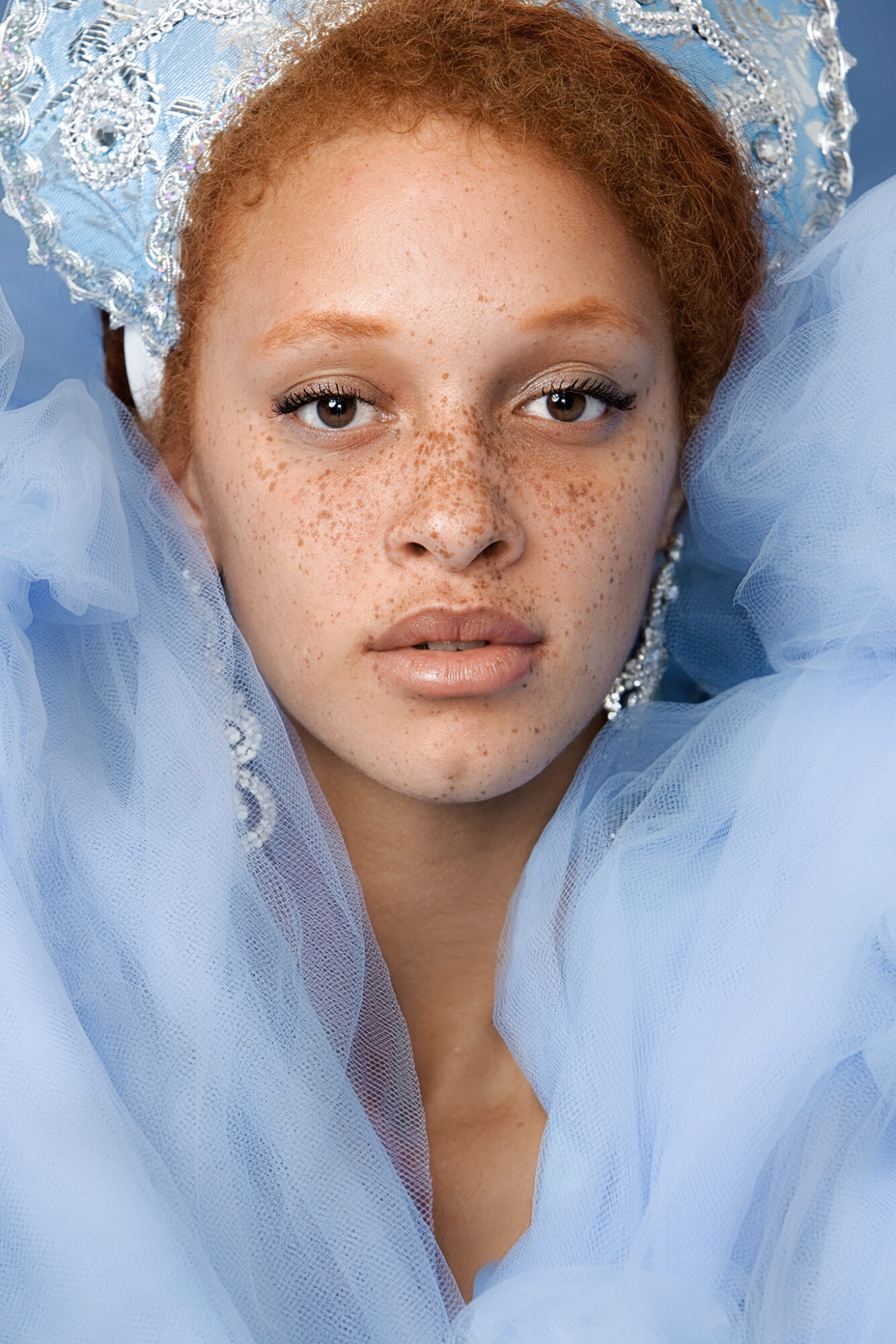 Close-up of a young woman with red hair, freckles, wearing a blue lace headpiece and matching dress, looking at the camera.