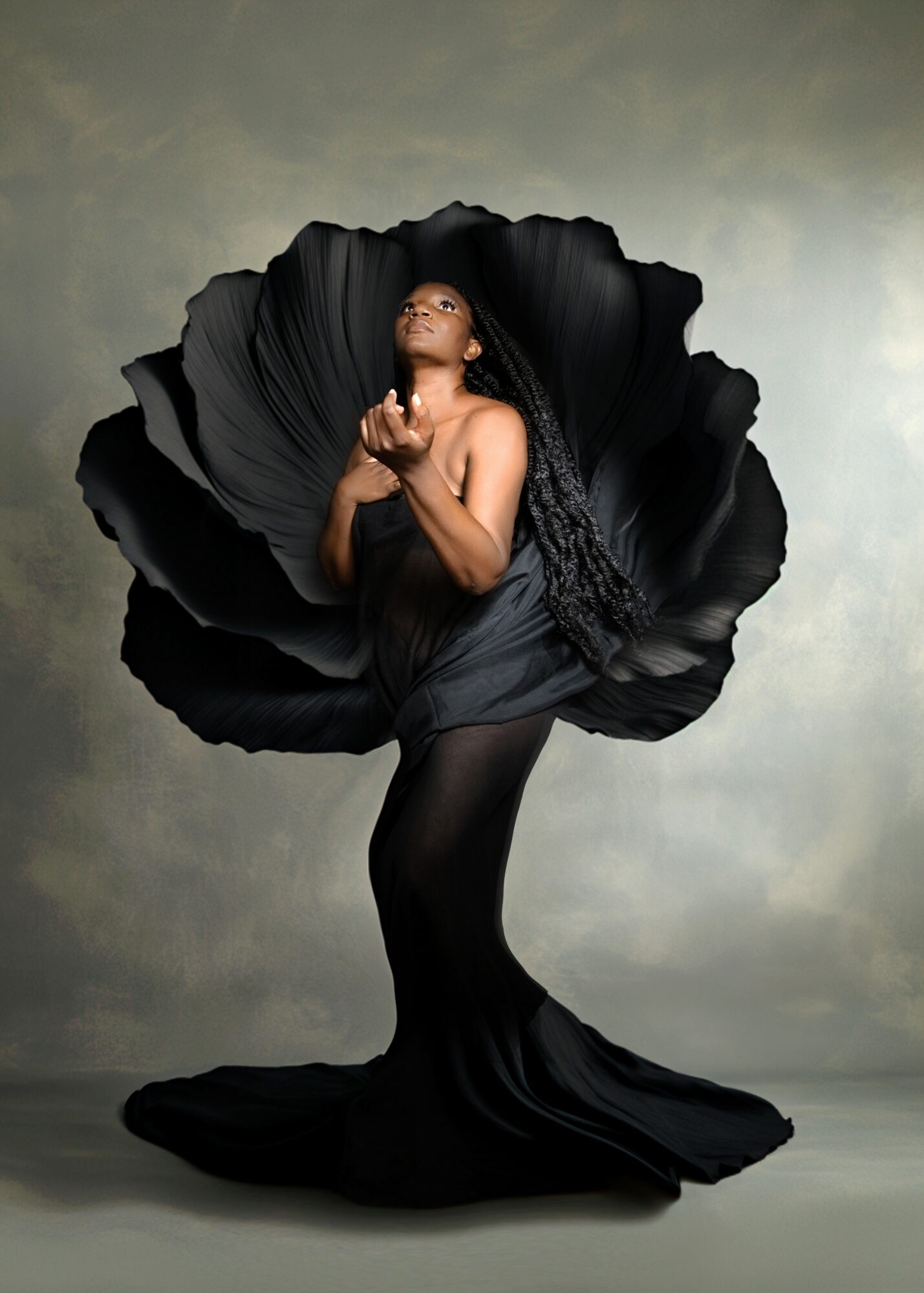 Woman in black dress with large flower-like fabric behind her, posing with hands near face, against a neutral background.