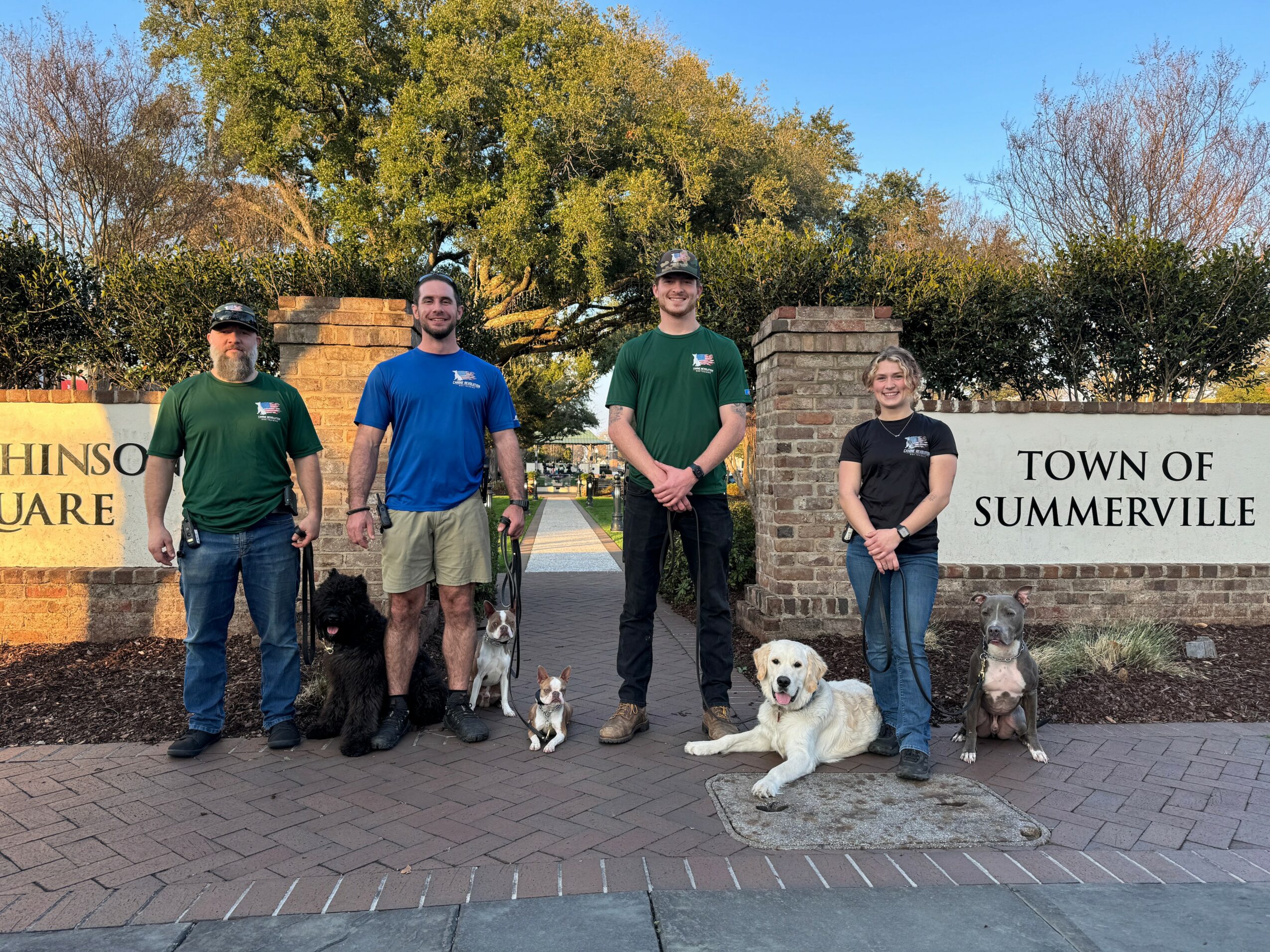 Four people stand outdoors in front of a brick sign, with dogs sitting or lying on the ground, trees, and a clear sky behind them.
