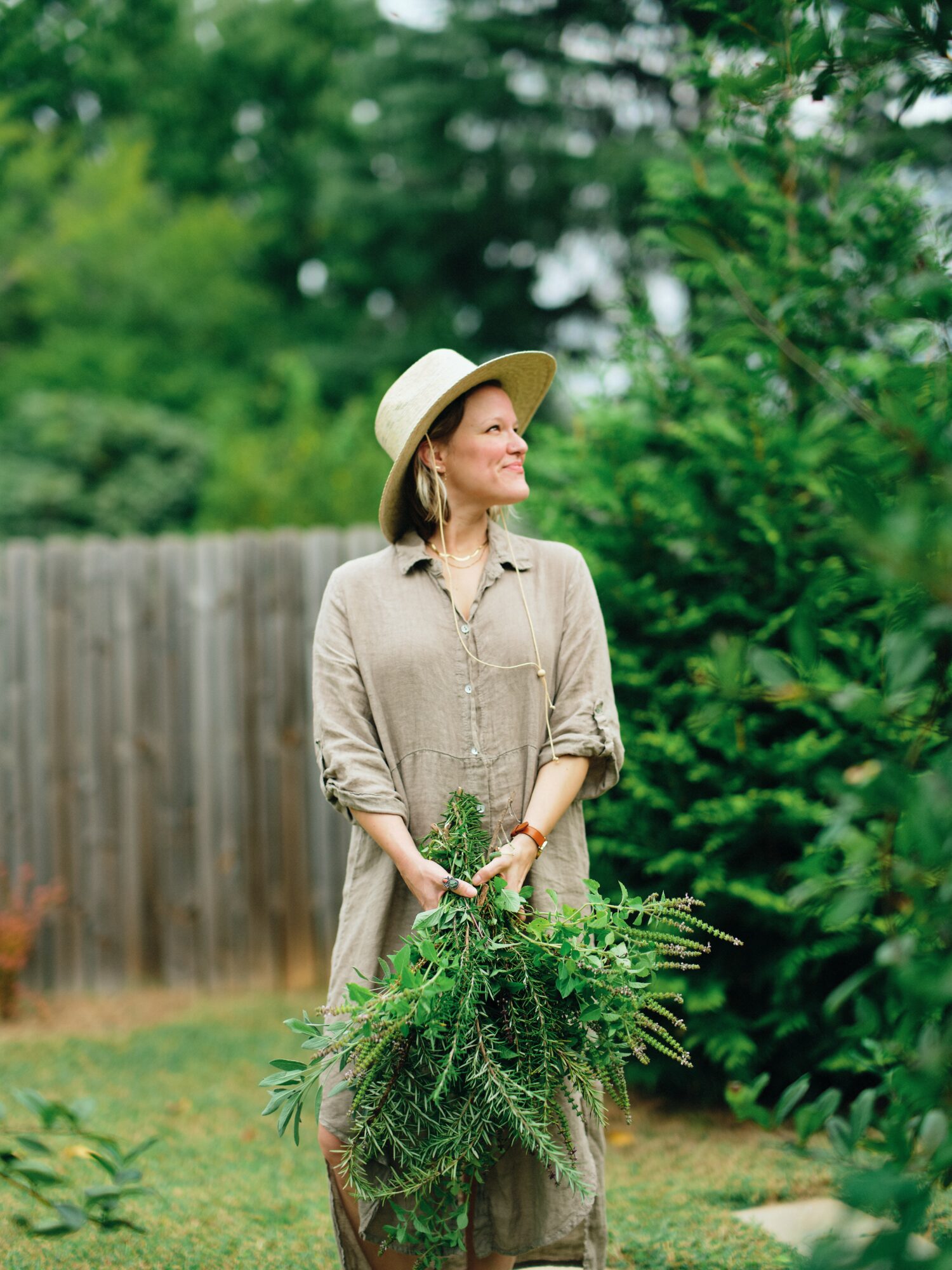 Woman in beige dress and wide-brimmed hat holding a large green plant outdoors, with trees and a wooden fence in background.