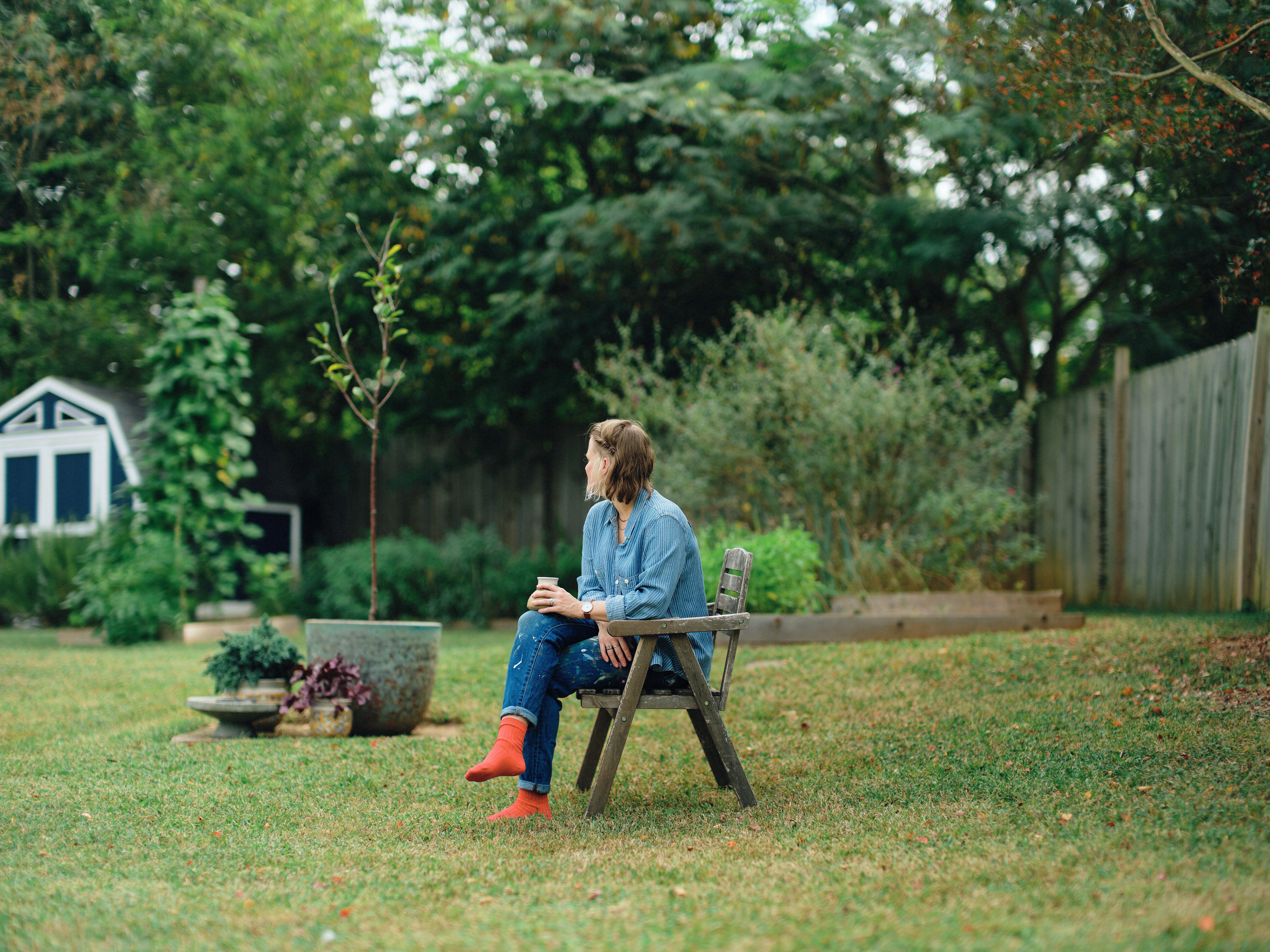 Person sitting on a wooden bench in a backyard with trees, plants, and a small shed, holding a cup.