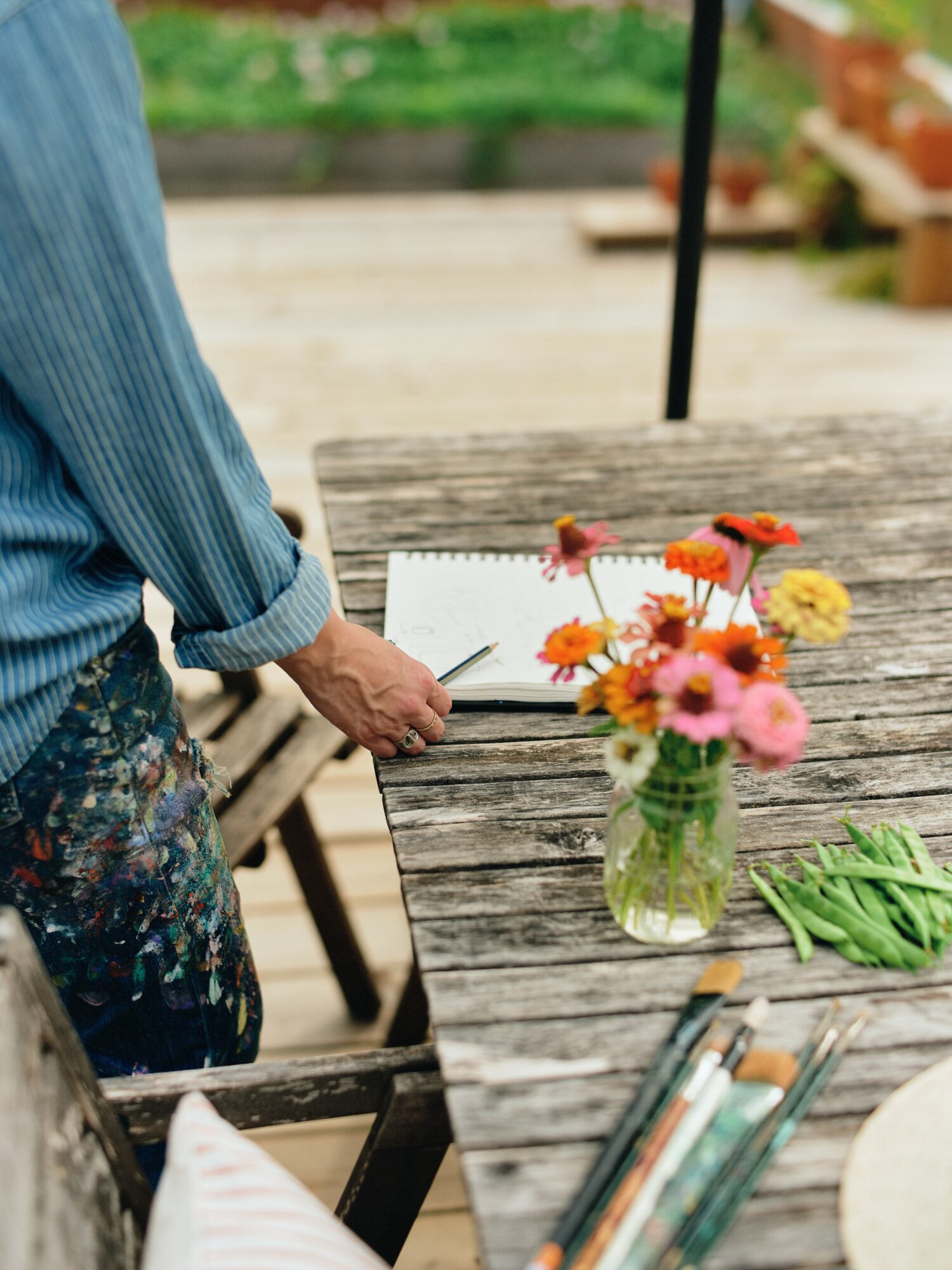 Person in blue shirt writing on paper at a wooden table with a vase of colorful flowers and green beans.