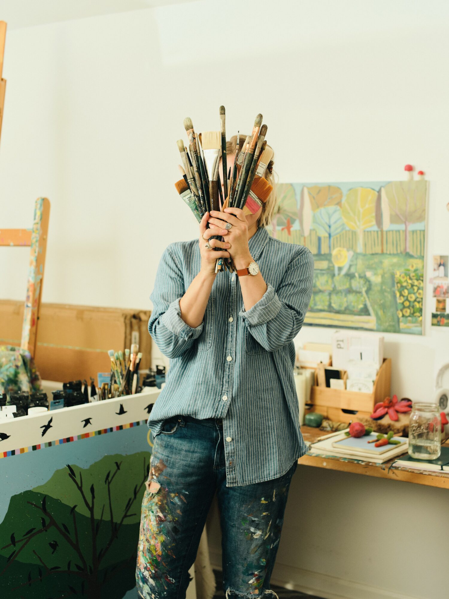 Person holding paintbrushes covering face, standing in art studio with paintings and supplies, casual clothing, jeans, and a striped shirt.