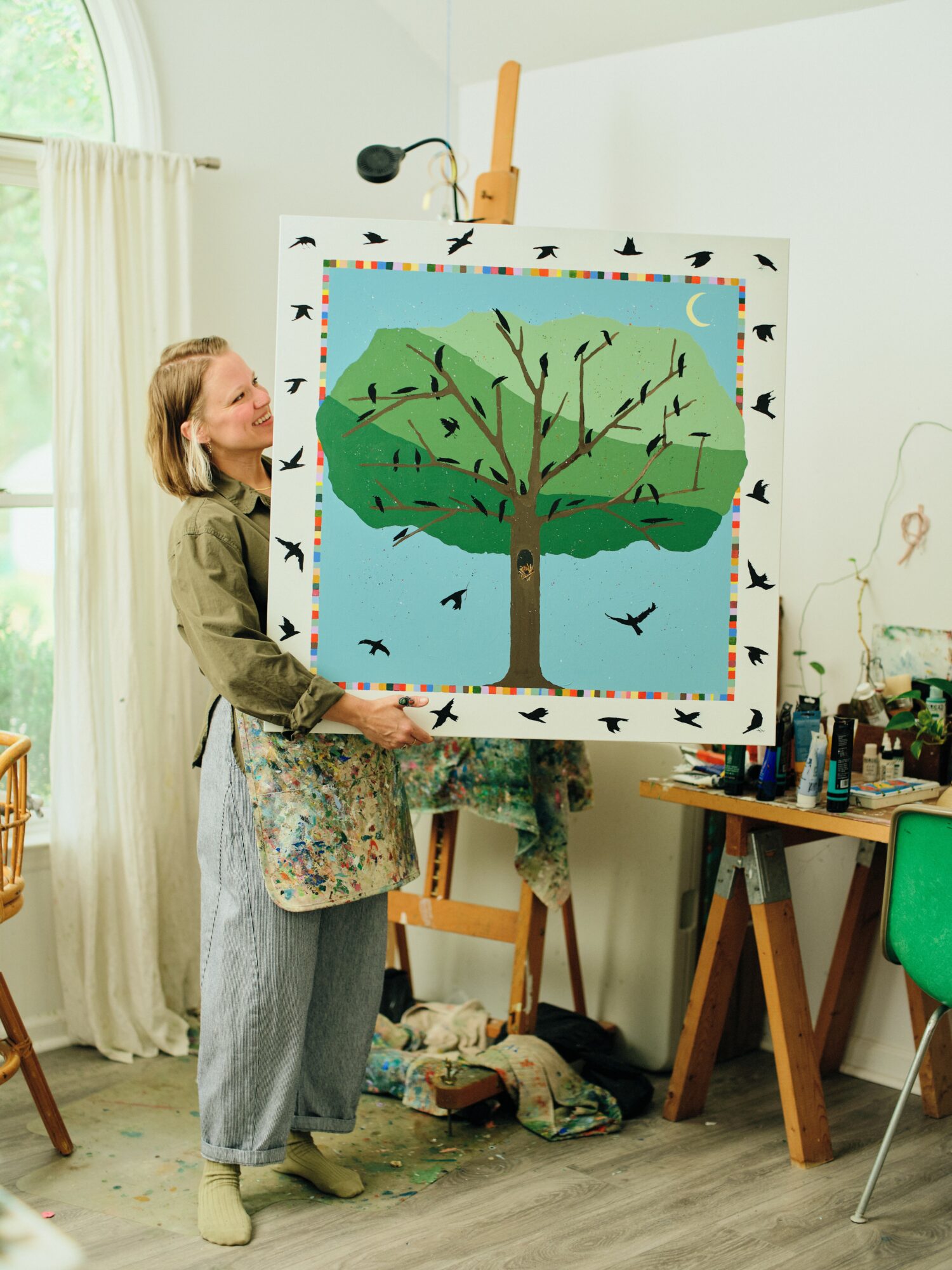 Person holding a colorful painting of a tree with birds, in an art studio with supplies and a window.