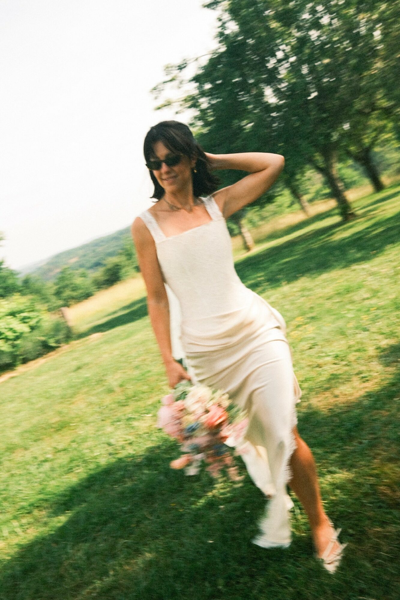 Woman in a white dress holding a bouquet outdoors, standing on grass with trees in the background.