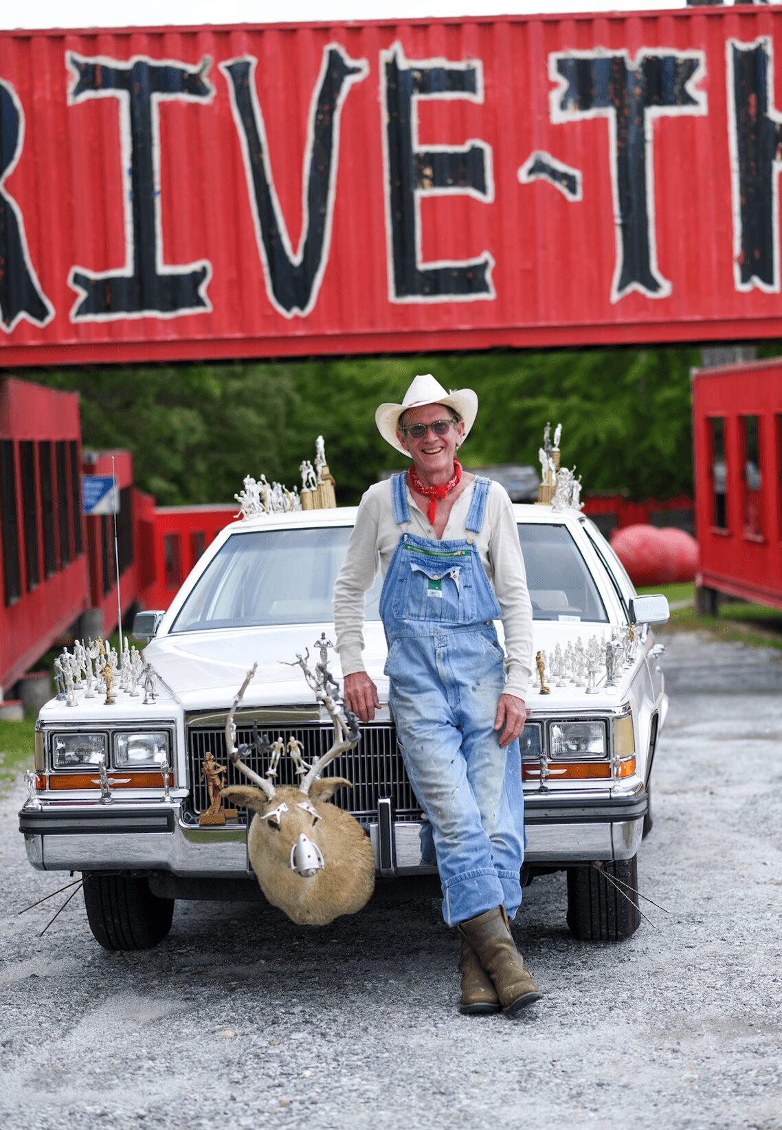 Person in cowboy hat and overalls standing in front of a decorated car with a reindeer figure, under a red sign with black text.