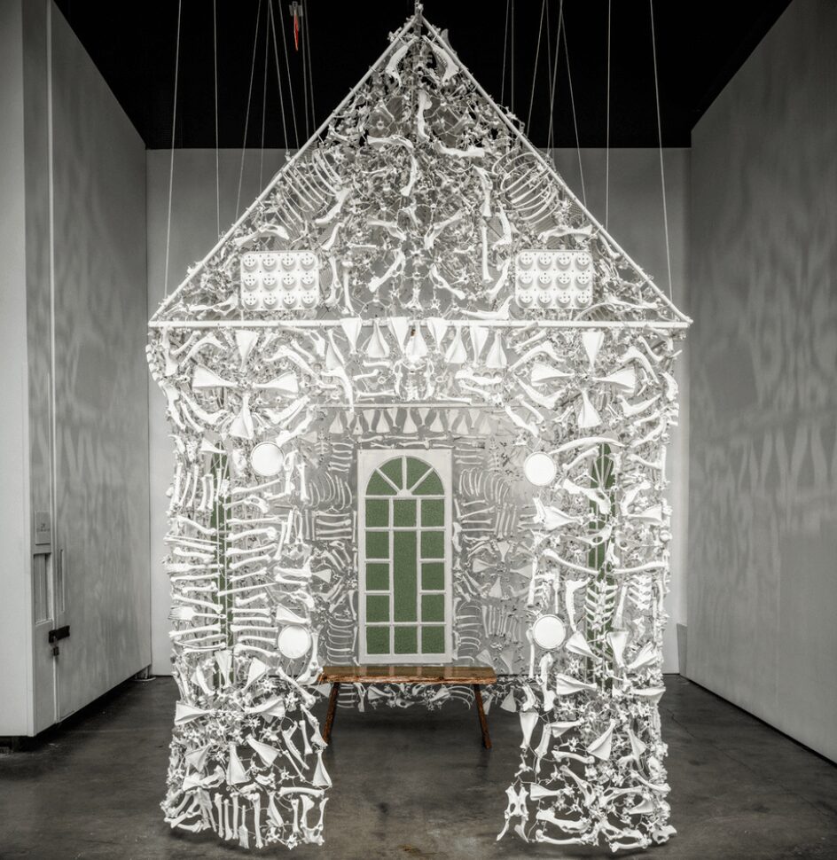 Intricate illuminated sculpture resembling a house with detailed patterns and a central door, displayed in a gallery setting.