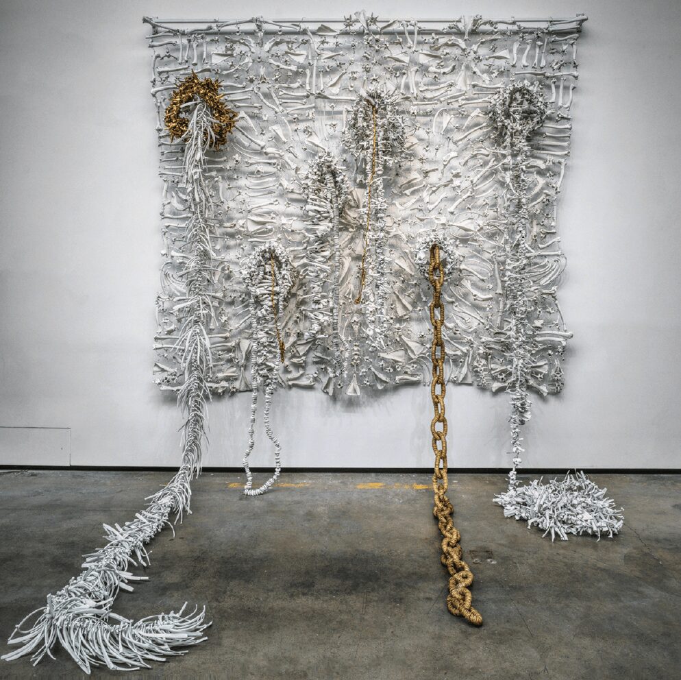 Mixed media art installation with metallic, chain, and textile elements on a wall, including a large chain hanging down.