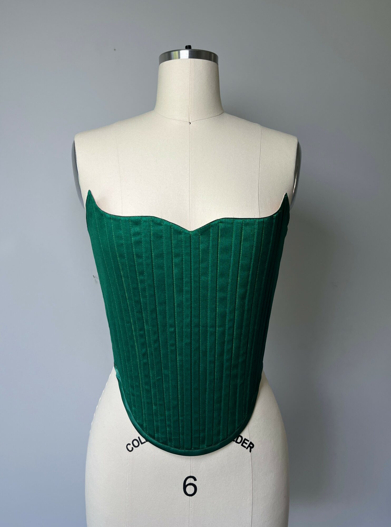 Green strapless corset on a dress form, with a scalloped top edge and vertical ribbing, against a plain background.