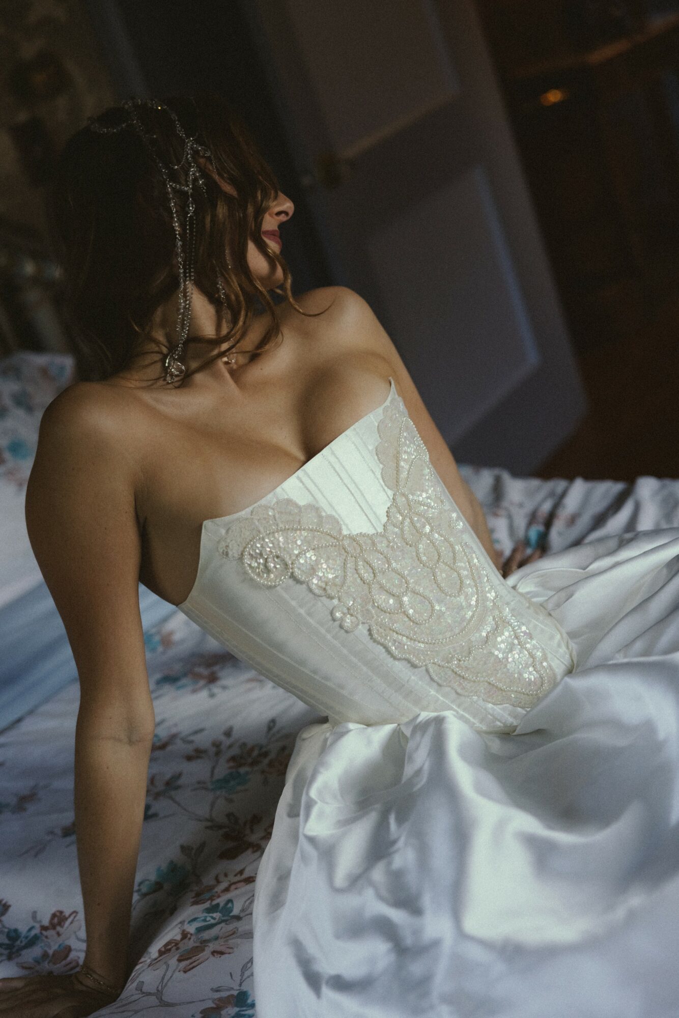 Woman in a white strapless dress sitting on a bed, facing left, with dark hair and a dimly lit background.