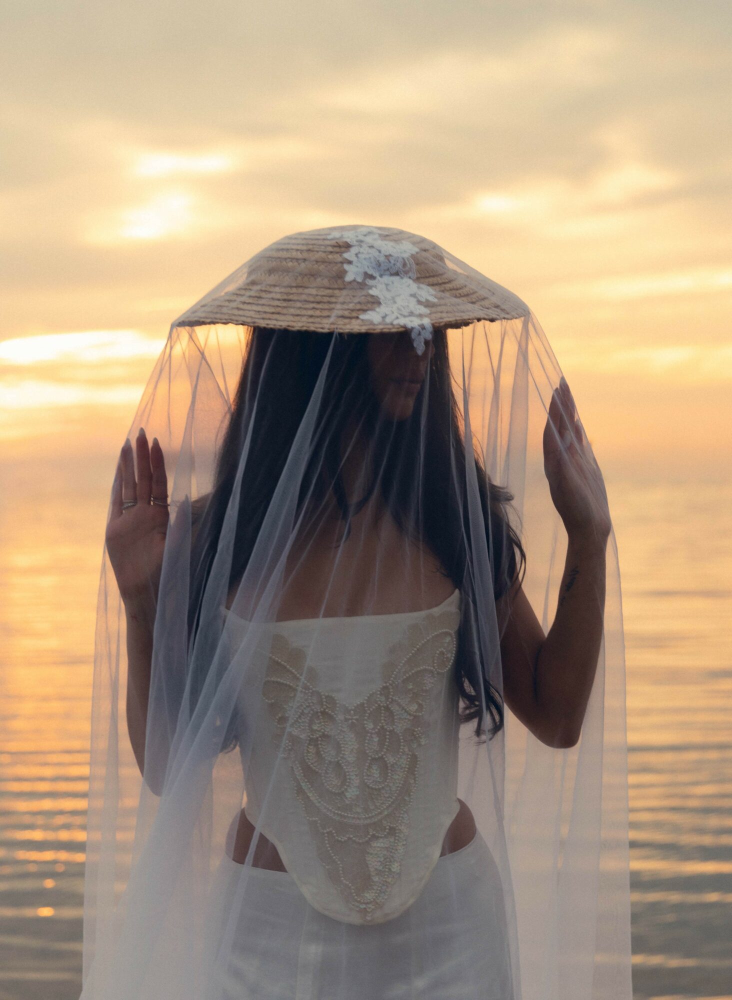 Person with long dark hair wearing a strapless dress and veil, holding a hat with flowers, standing by water at sunset.