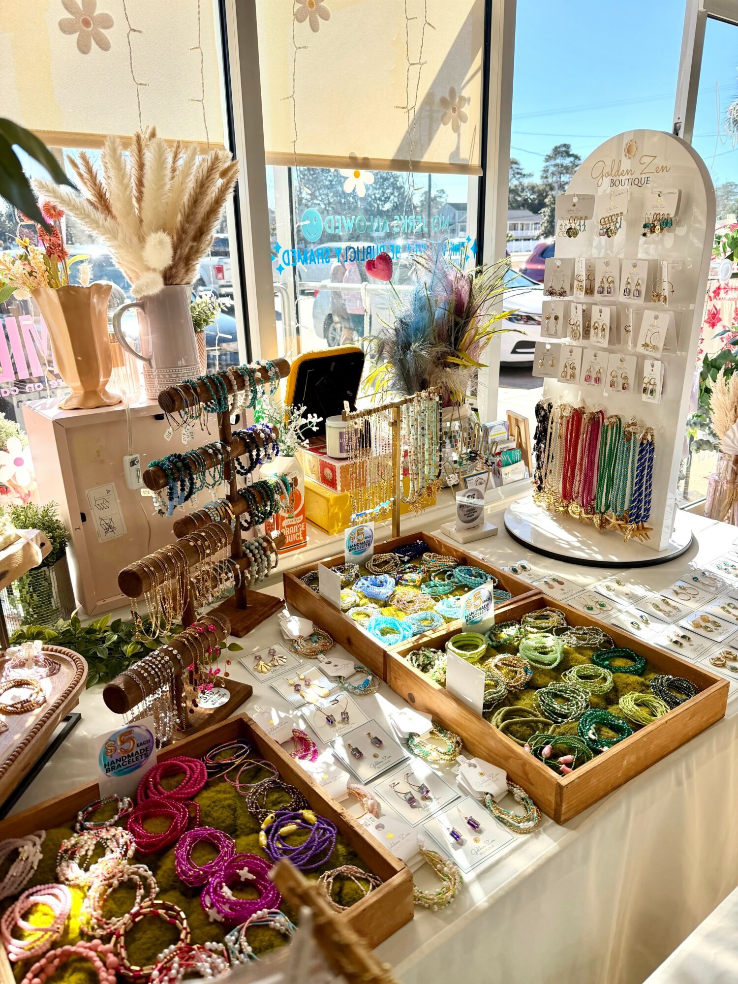Display of jewelry, accessories, and decorative items on a table near a window, with sunlight and outdoor view.