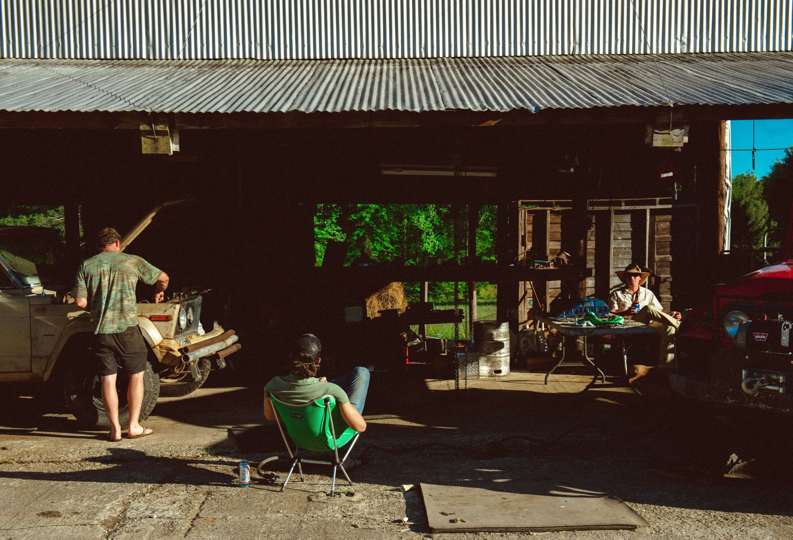 People gather outside a rustic building with a corrugated metal roof, some sitting and others standing near parked cars.