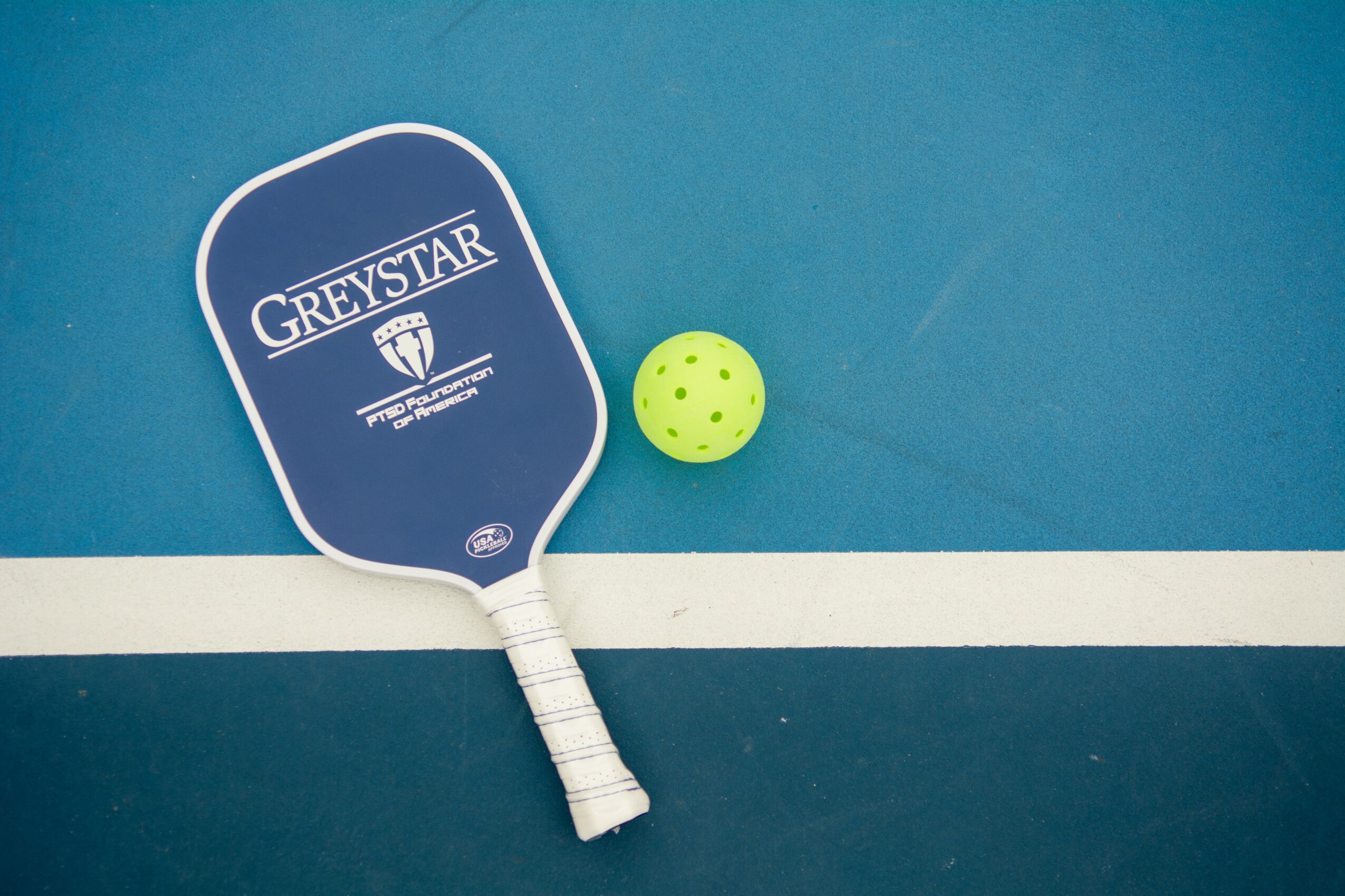 Padel racket, yellow ball, and court line on blue surface, viewed from above.