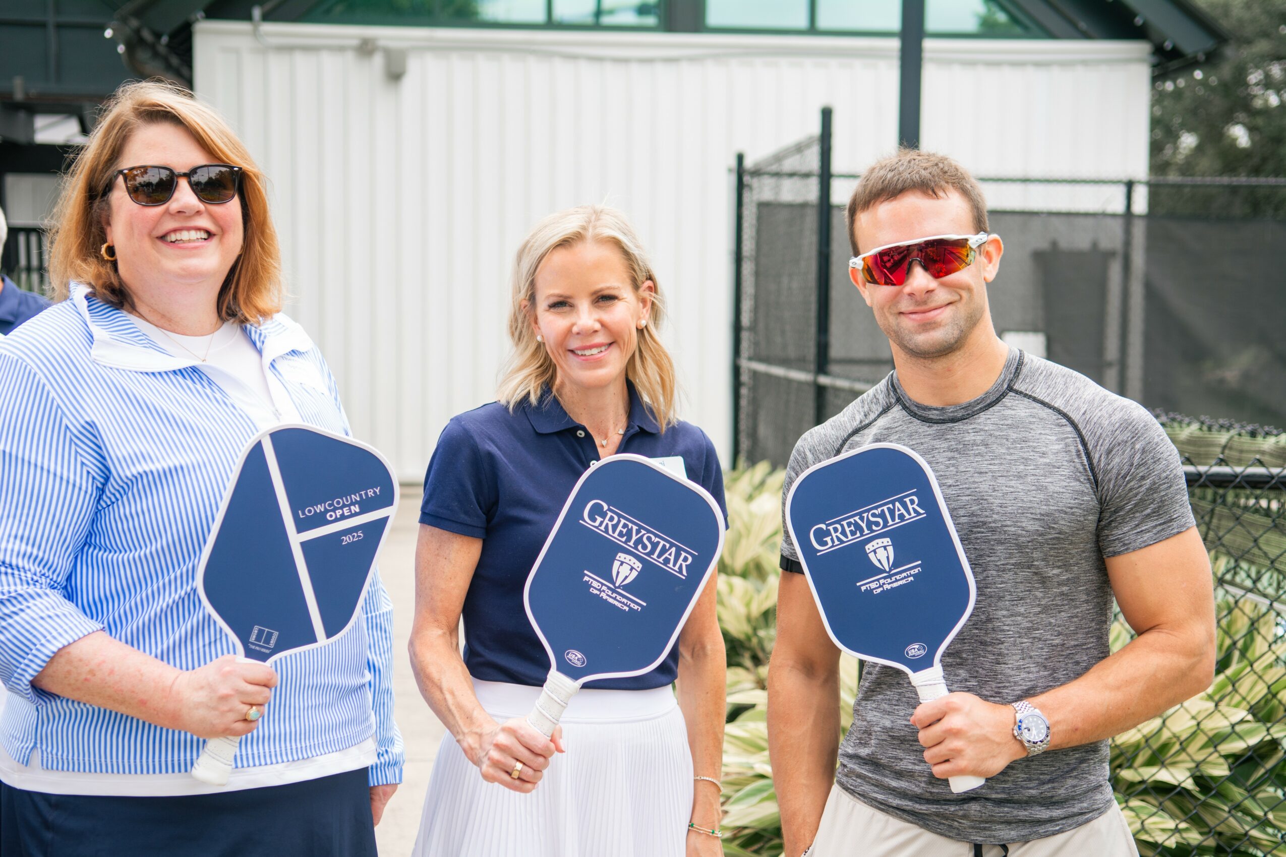 Three people outdoors holding blue paddle-shaped signs, smiling, with a fence and building in background.