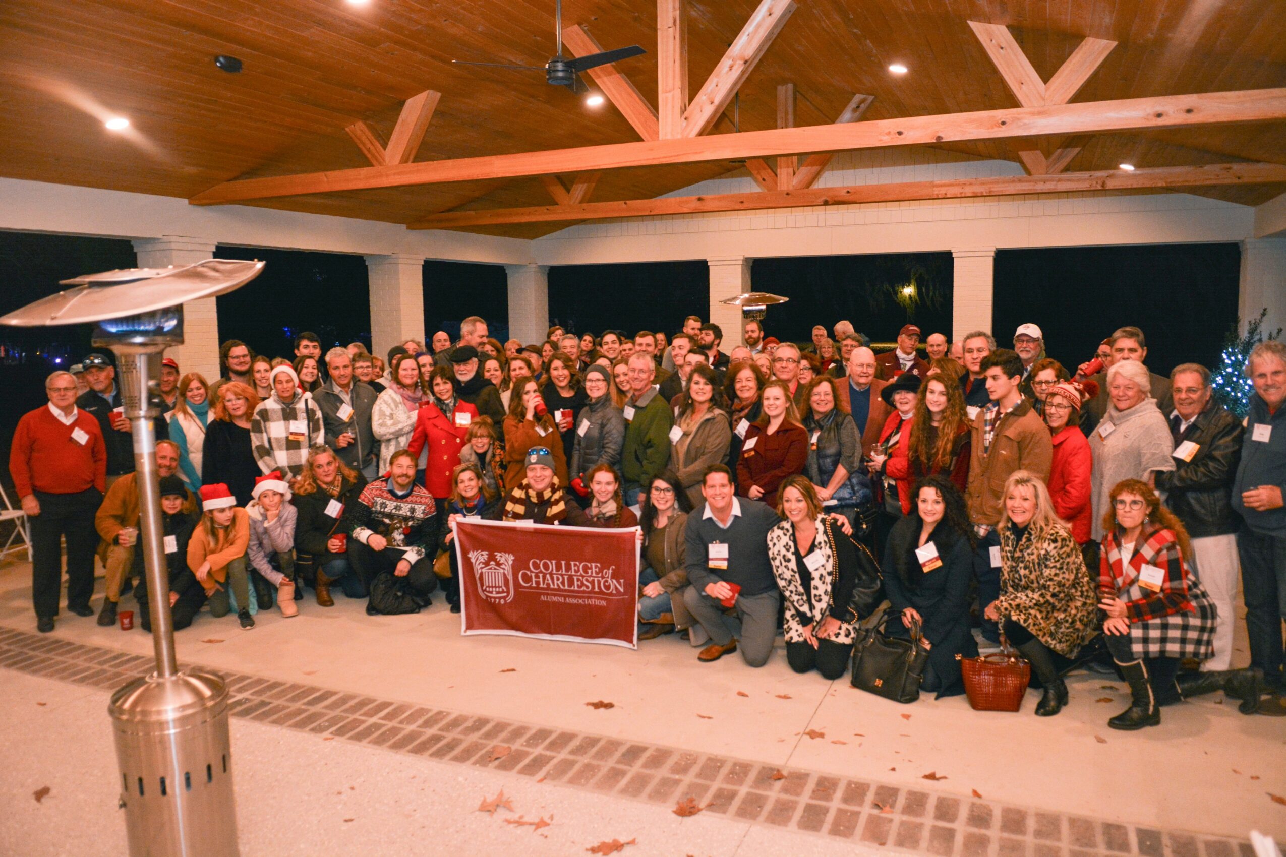 Large group of people gathered indoors, posing for a photo with a banner, in a room with wooden ceiling beams.