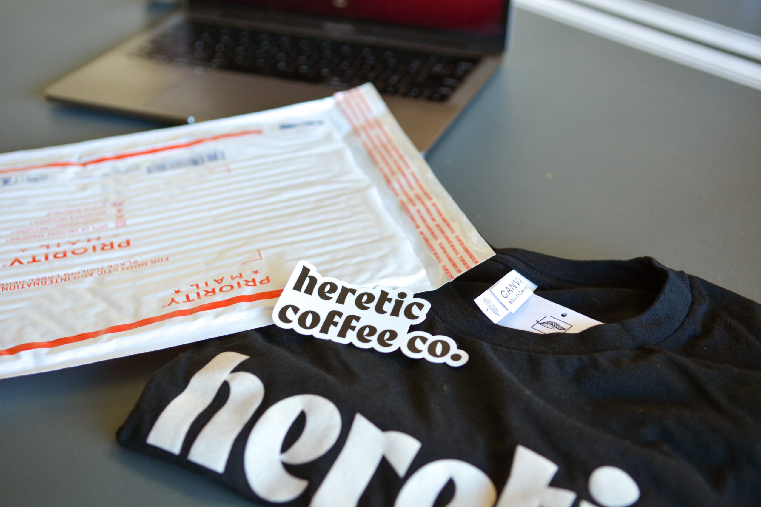 Black T-shirt with white text reading 'heretic coffee co.' on a table, with a package and a laptop in the background.