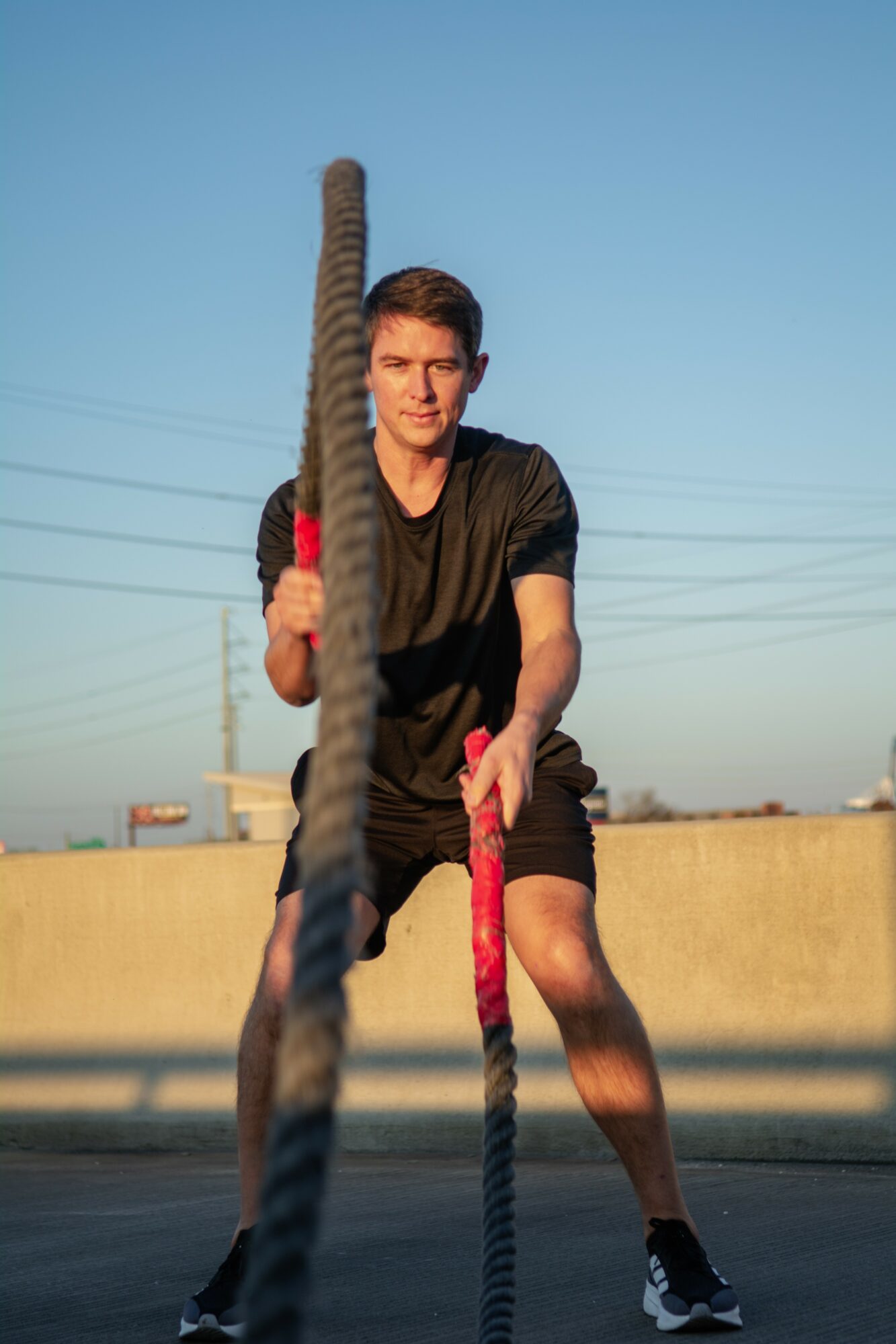 Young man holding battle ropes outdoors on a rooftop, facing the camera, with a clear sky background.