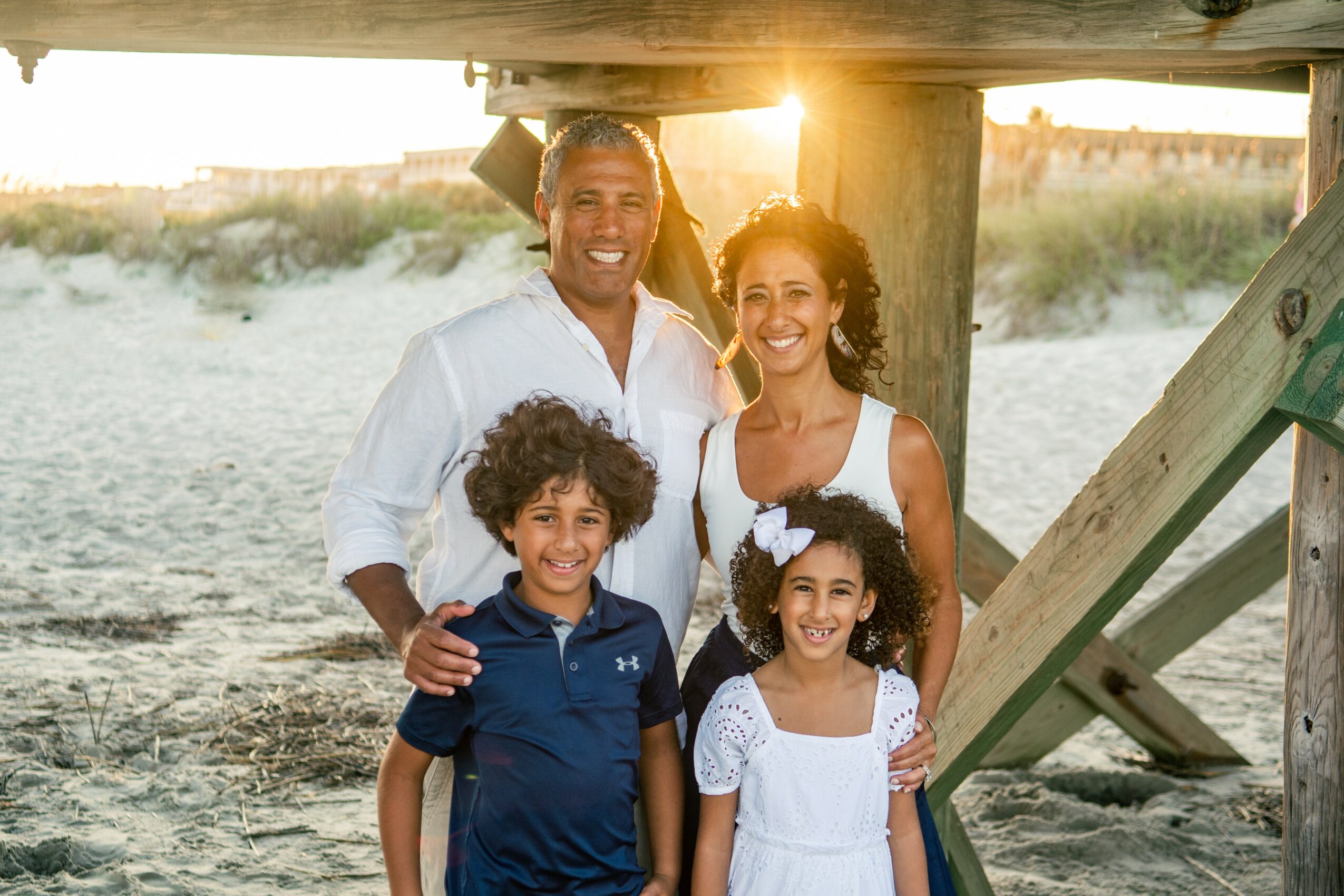 Family of four smiling under a wooden structure on a beach at sunset, with sand and grass in the background.