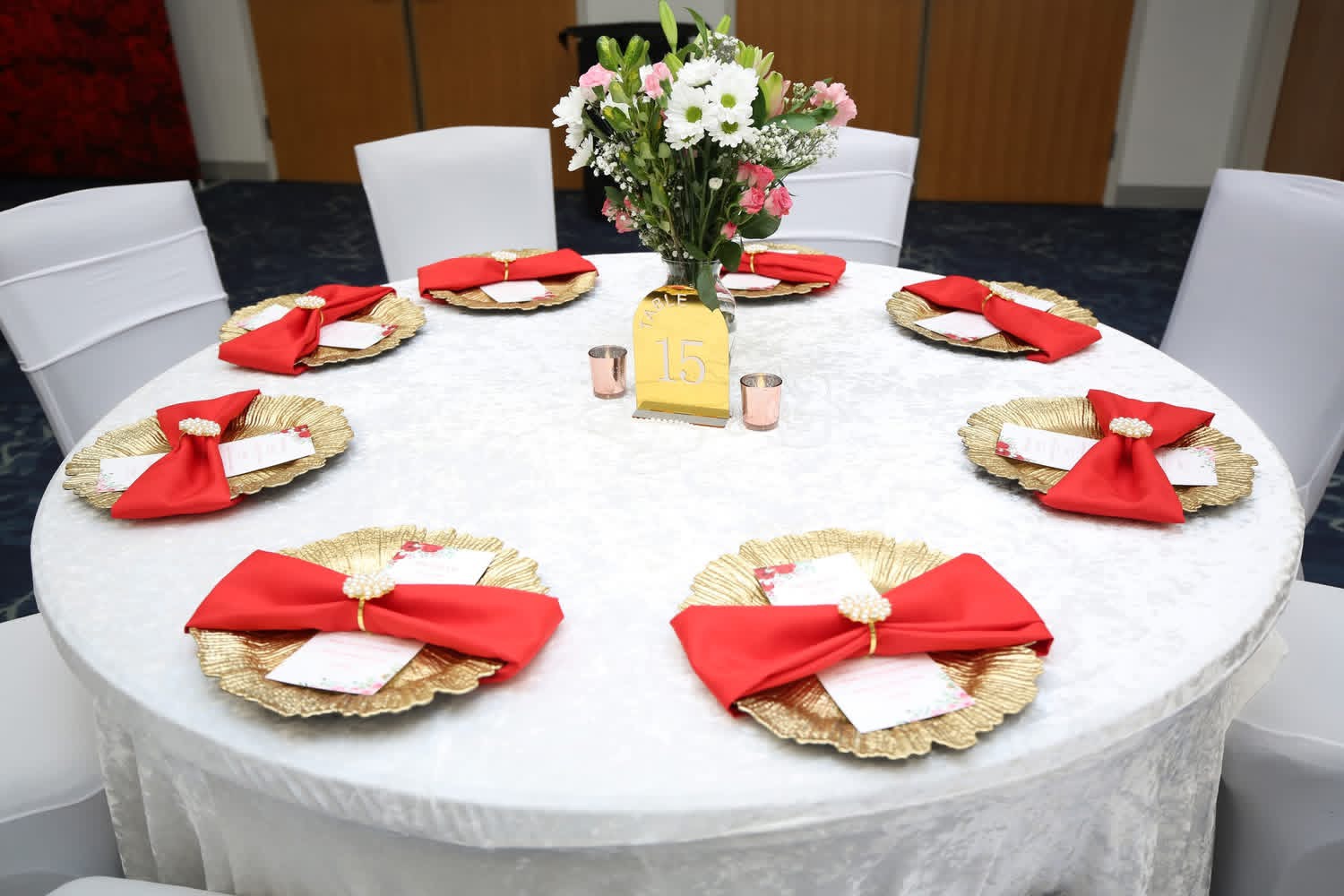 Round table with white tablecloth, six place settings with red napkins, and a floral centerpiece with white and pink flowers.