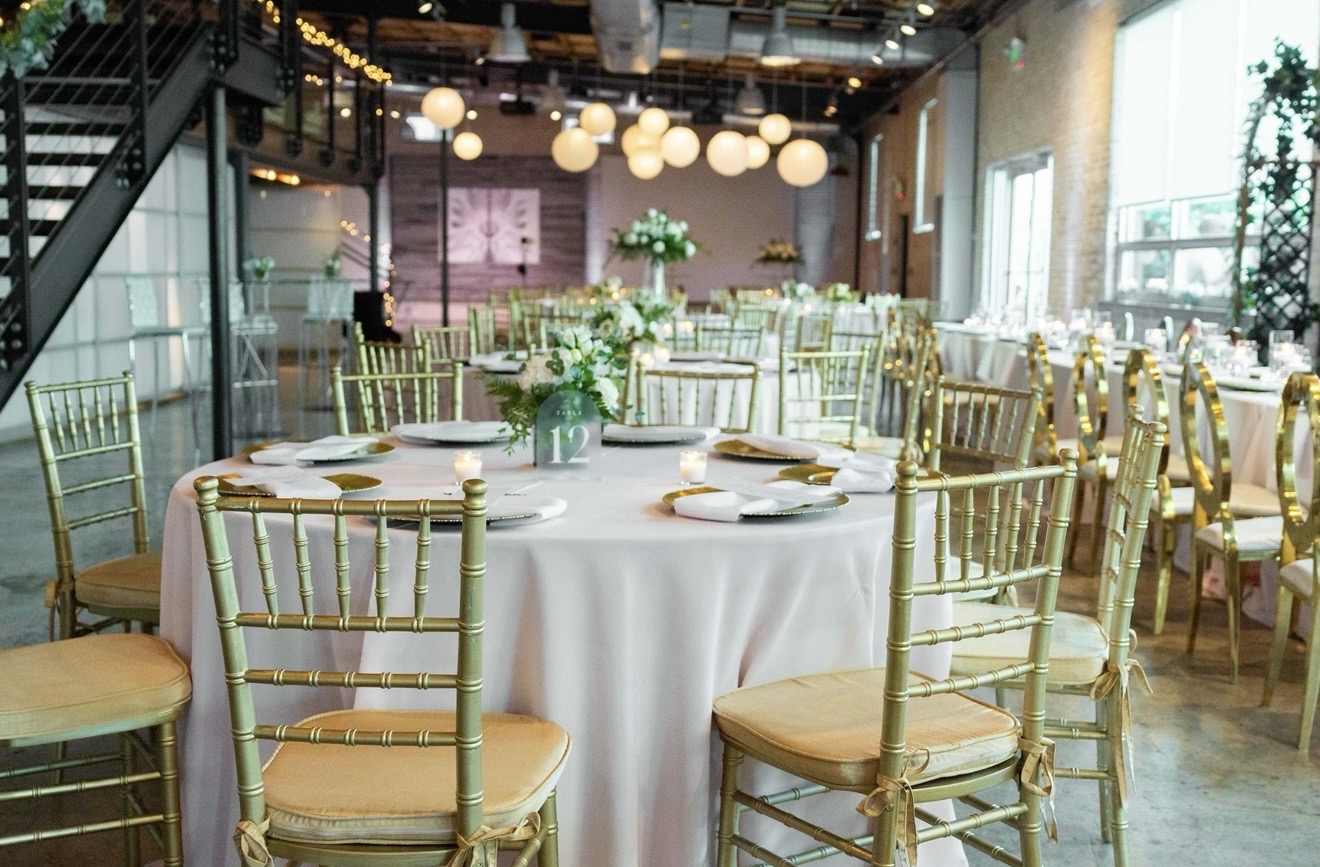 Elegant event space with round tables, gold chairs, white tablecloths, and floral centerpieces, with large windows and modern lighting.