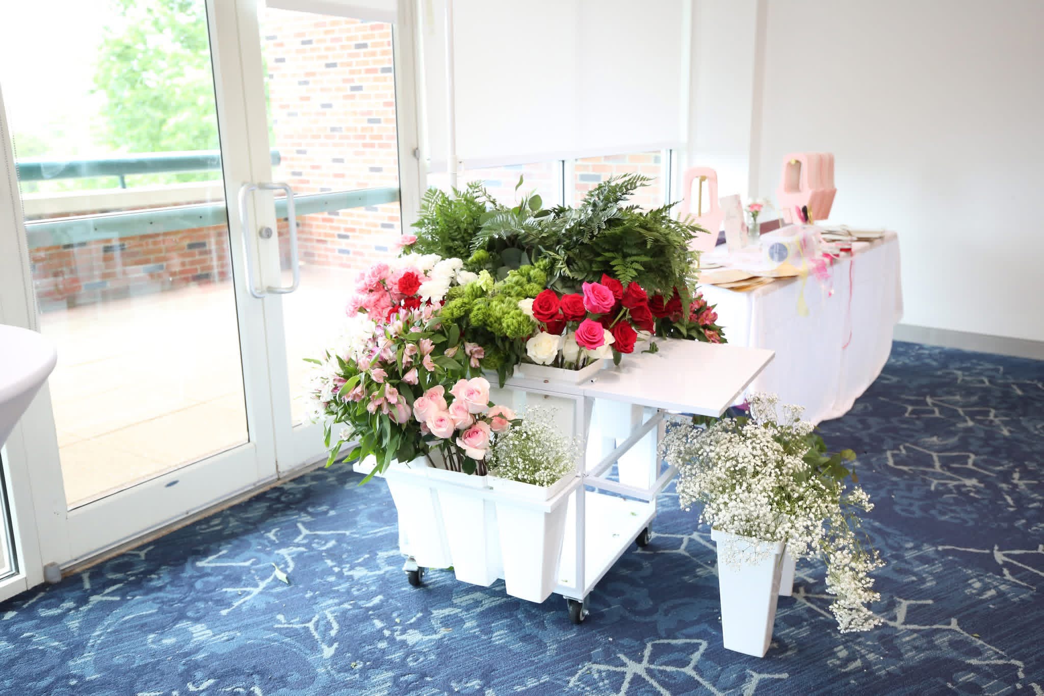 Table with pink and white flowers, greenery, and a small white vase with baby's breath, near glass door, blue carpeted floor.