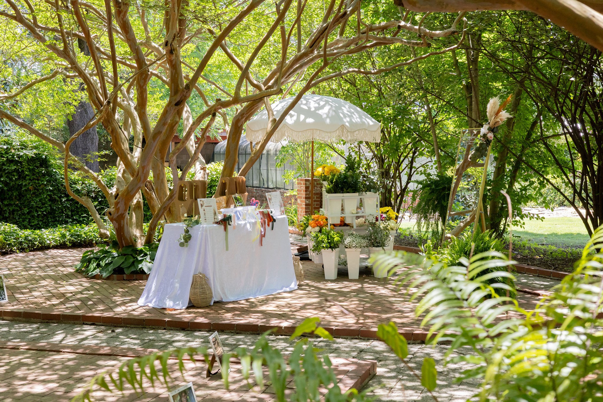 Outdoor garden scene with tables, chairs, umbrella, and potted plants under trees, surrounded by greenery and sunlight.