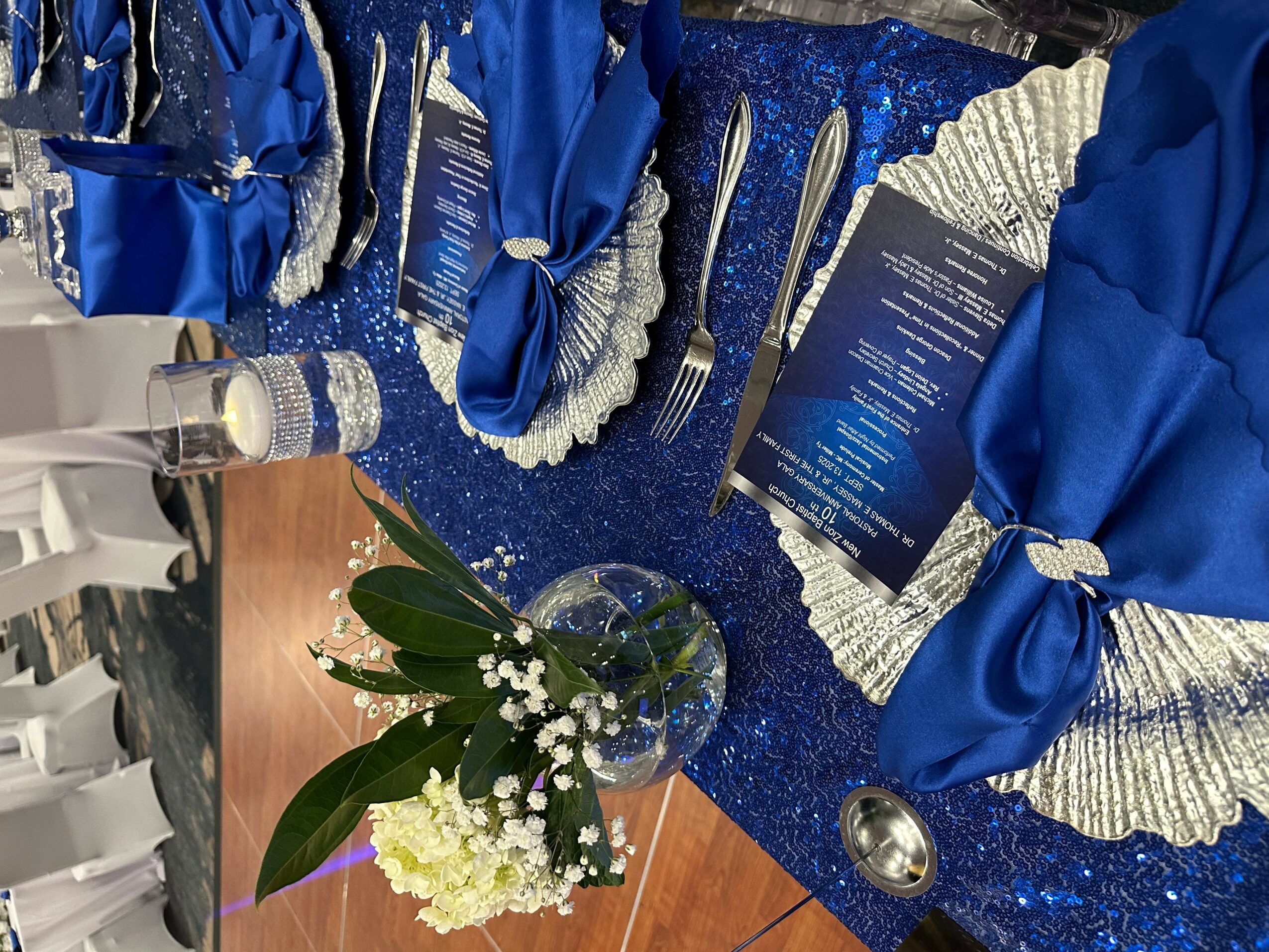 Table setting with blue and silver napkins, menus, and a floral centerpiece with white flowers and green leaves.