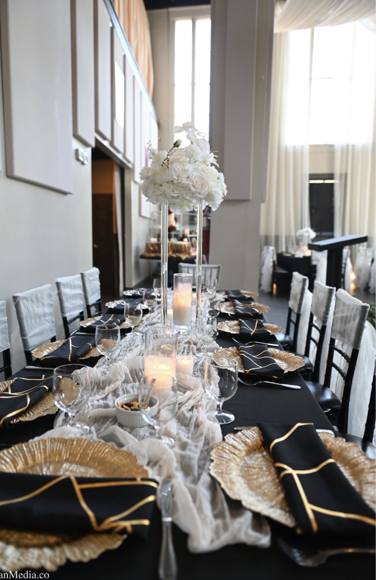 Elegant dining table set with black and gold napkins, white floral centerpiece, candles, and decorative elements in a bright room.