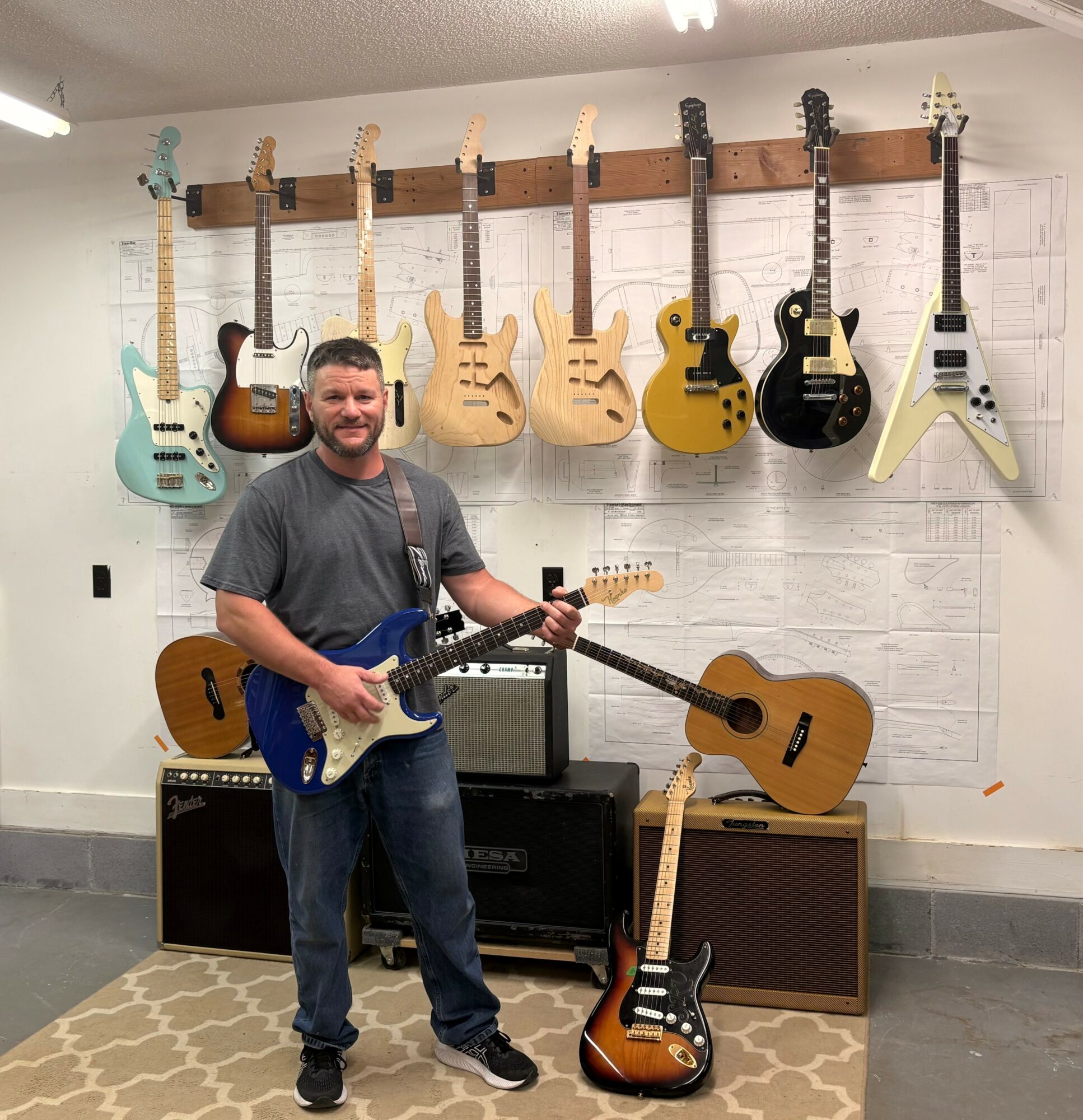 Man holding a blue electric guitar in a room with multiple guitars hanging on the wall behind him.