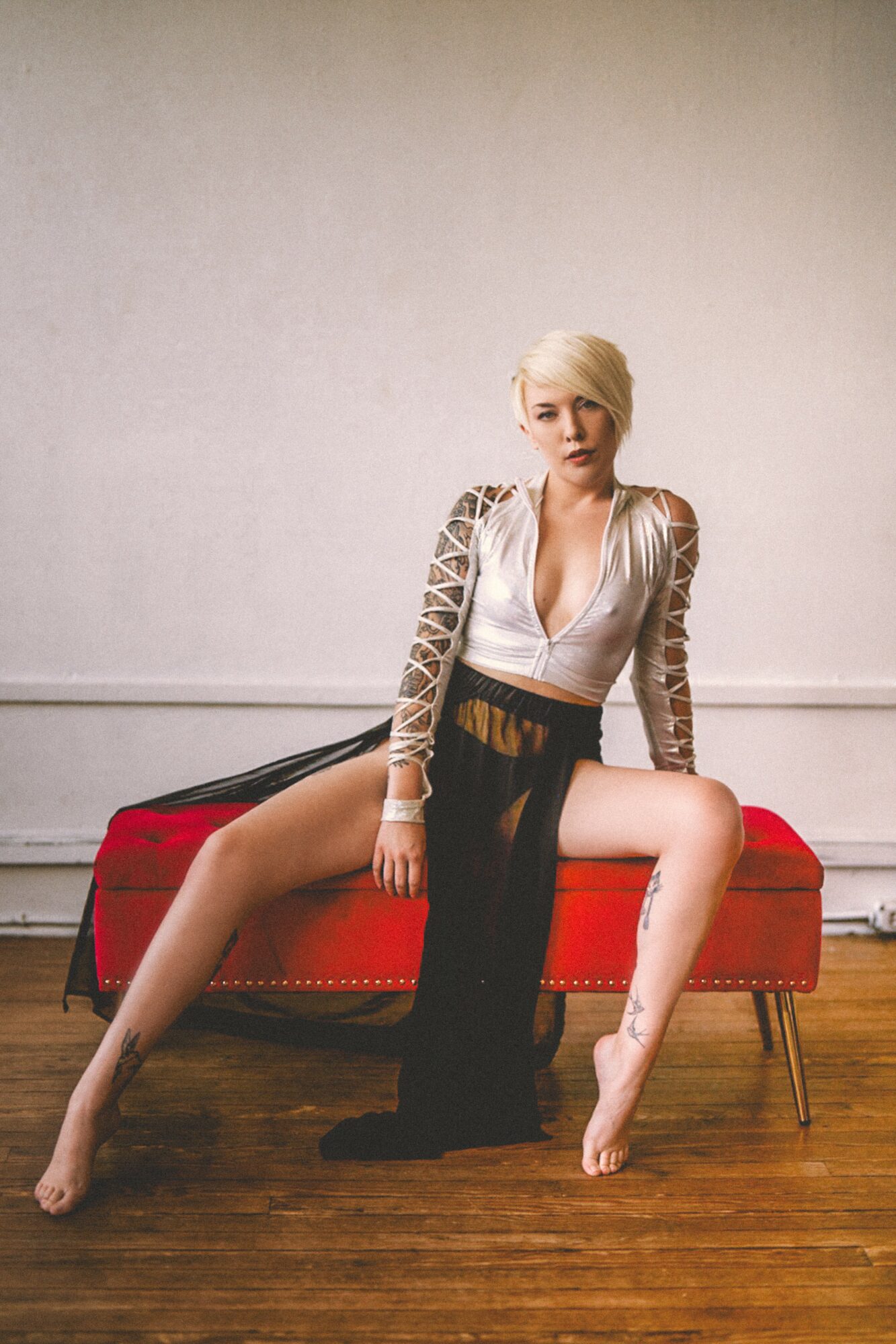 Woman with short blonde hair sitting on a red bench, legs spread, in a room with plain wall and wooden floor.