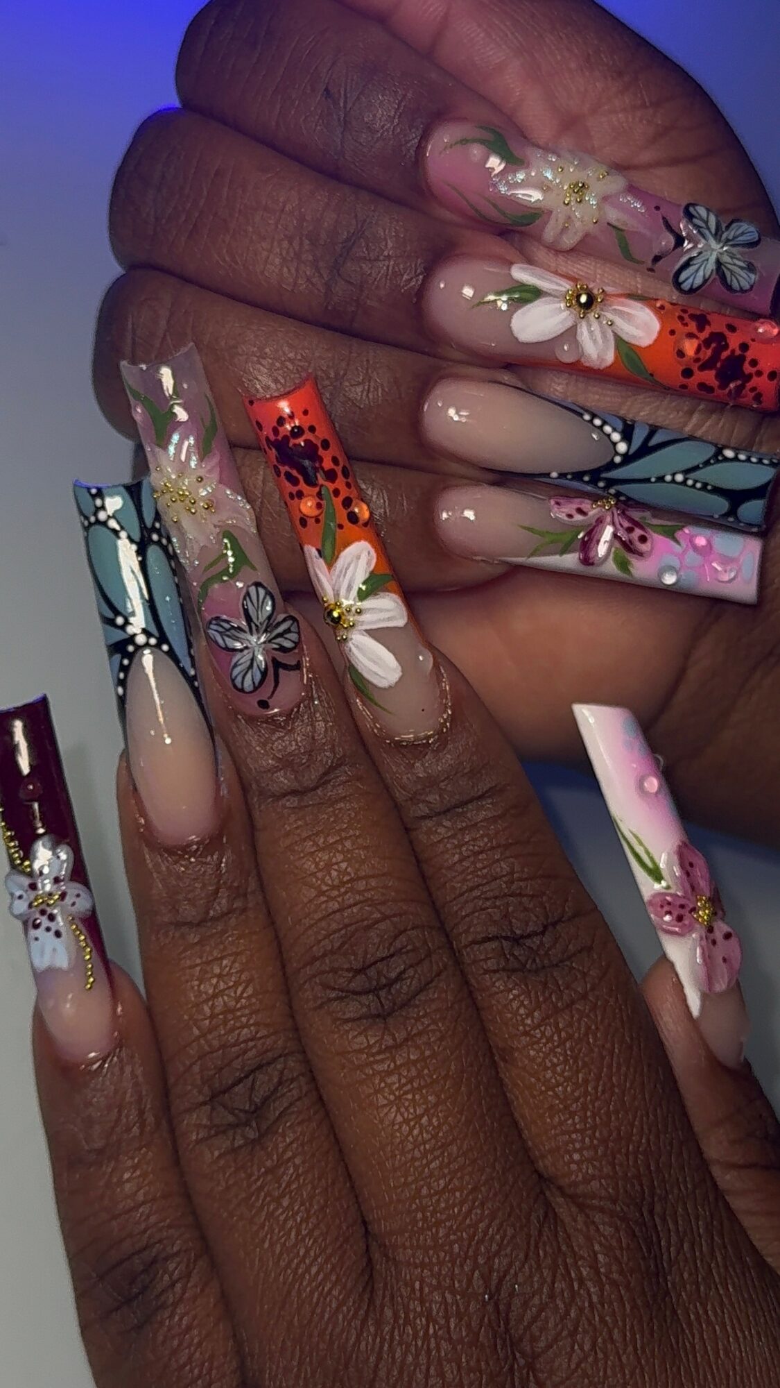 Close-up of hands with long, decorated nails featuring floral and butterfly designs, in various colors and patterns.