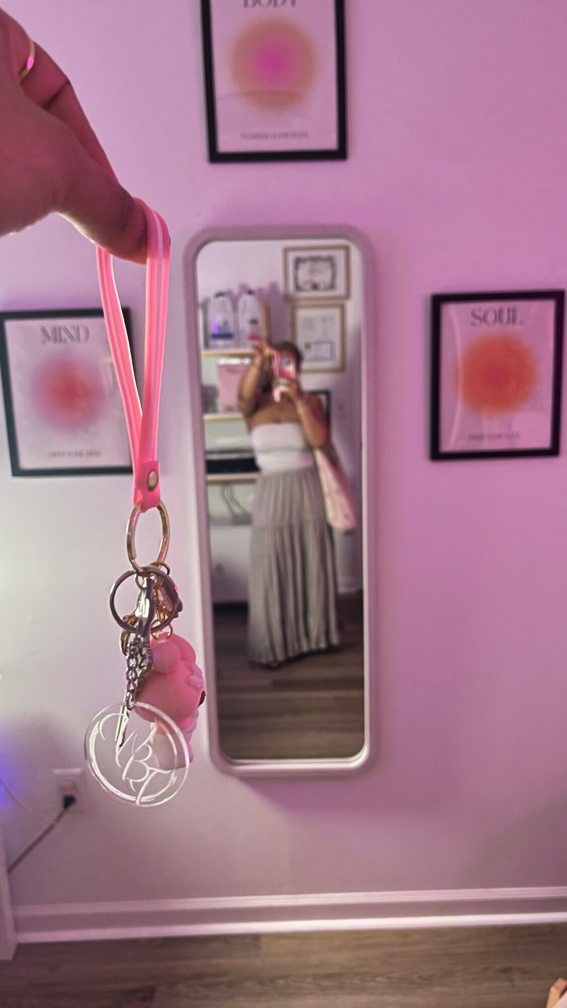 Person holding pink lanyard with keychain, mirror reflects woman in striped skirt, pink wall with framed art, and wooden floor.