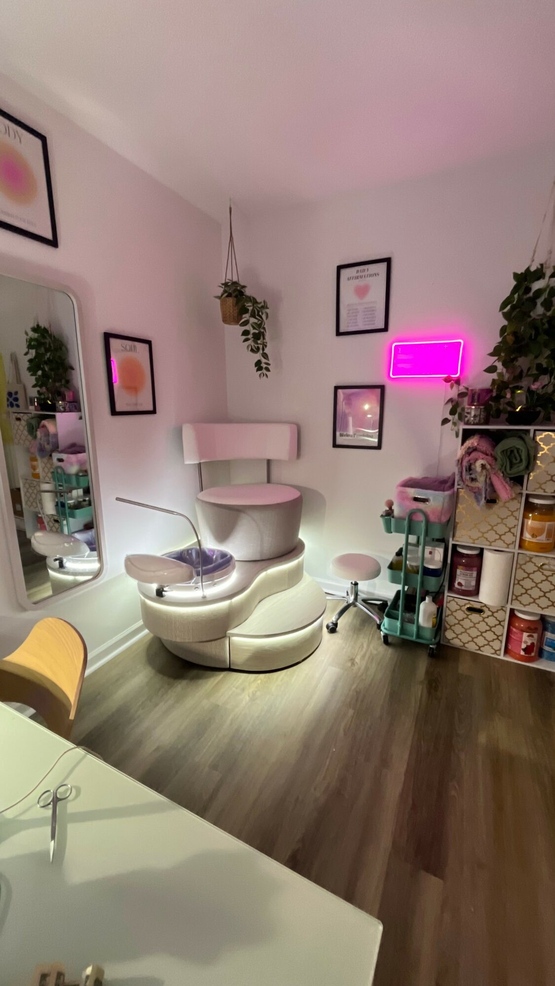 Room with white chair, wall art, pink neon sign, plants, and shelves with storage boxes, wooden floor, and a mirror.