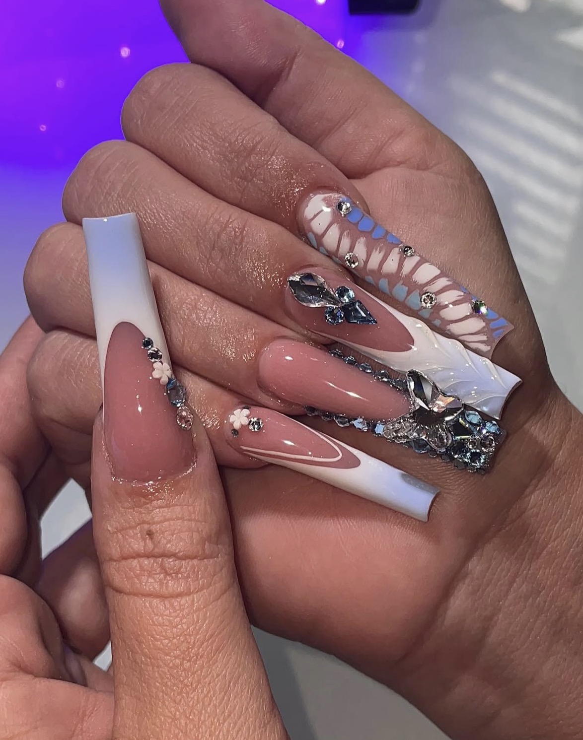 Close-up of hands with long, decorated acrylic nails featuring pink, white, and silver designs, rhinestones, and patterns.