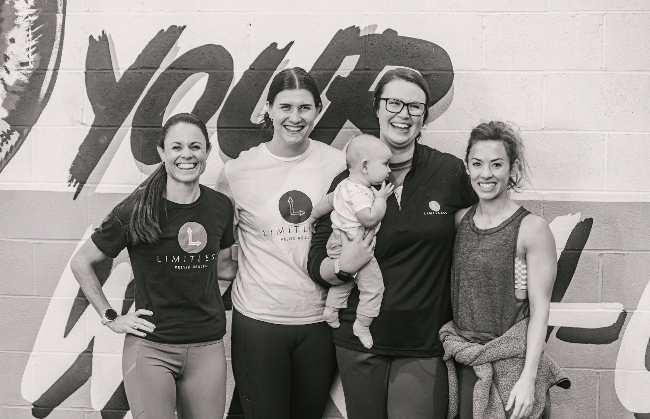 Four women and a baby standing in front of a wall with large text, smiling, in a group photo.