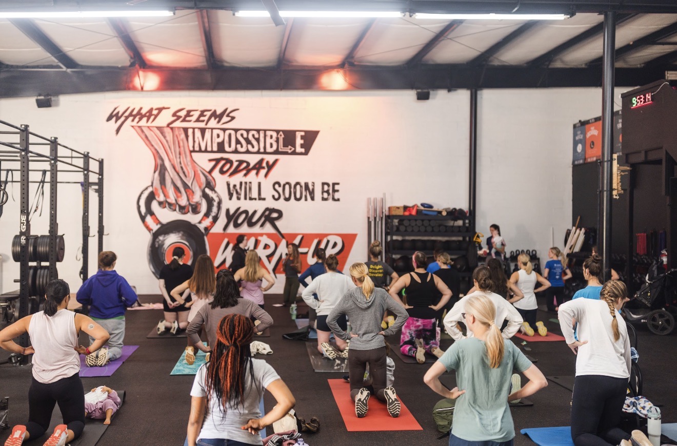 People participating in a fitness class in a gym with a motivational wall message, some on mats, others standing or stretching.