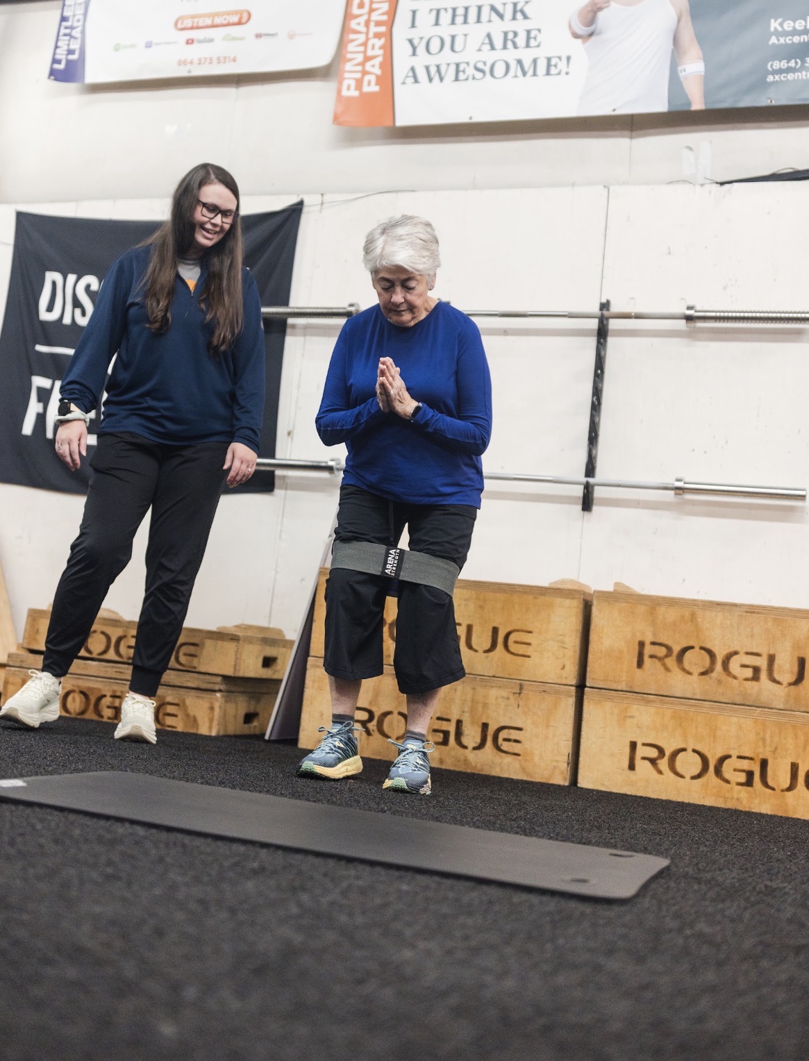 Two women in a gym, one with gray hair and the other with long dark hair, standing near a black mat and wooden boxes.