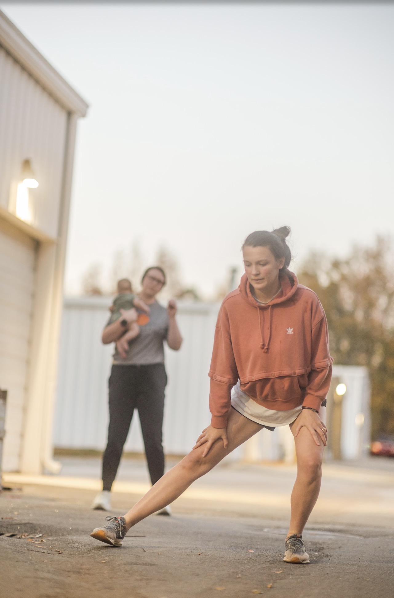 Person stretching outdoors with another person holding a child in the background, during daytime.