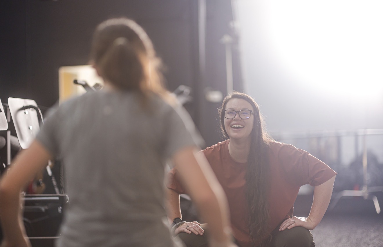 Two women smiling and talking, one with long hair and glasses, in a bright, indoor setting.