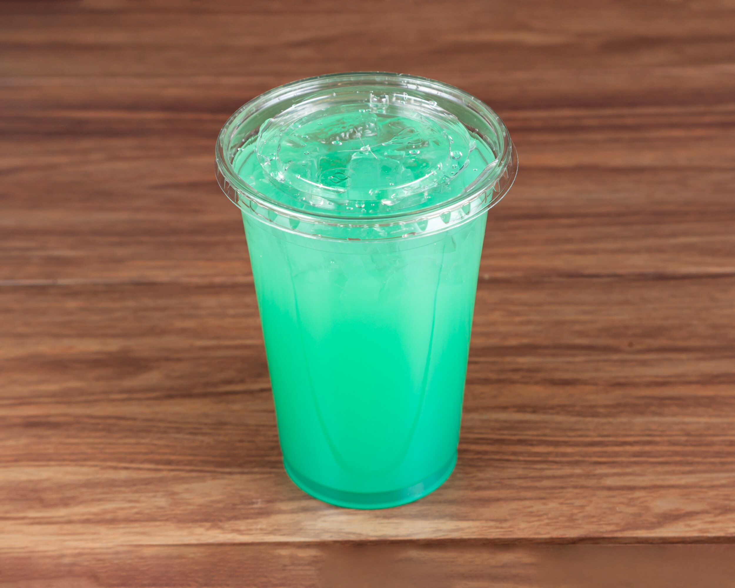 A clear plastic cup filled with a bright blue-green beverage and ice, placed on a wooden surface.