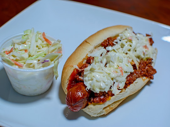 Hot dog with chili, cheese, and coleslaw on a bun, served with a side of coleslaw in a cup, on a blue plate.